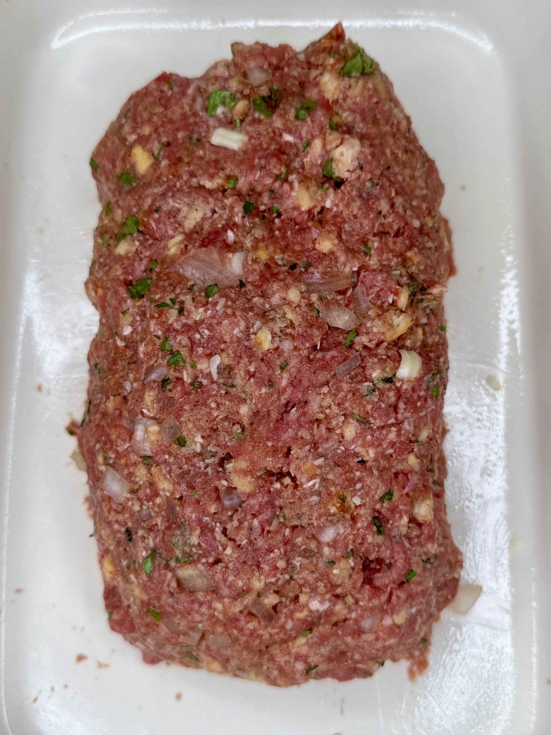 A raw meatloaf mixture shaped into a rectangular loaf, containing ground meat, chopped onions, herbs, and seasonings, placed on a white tray.