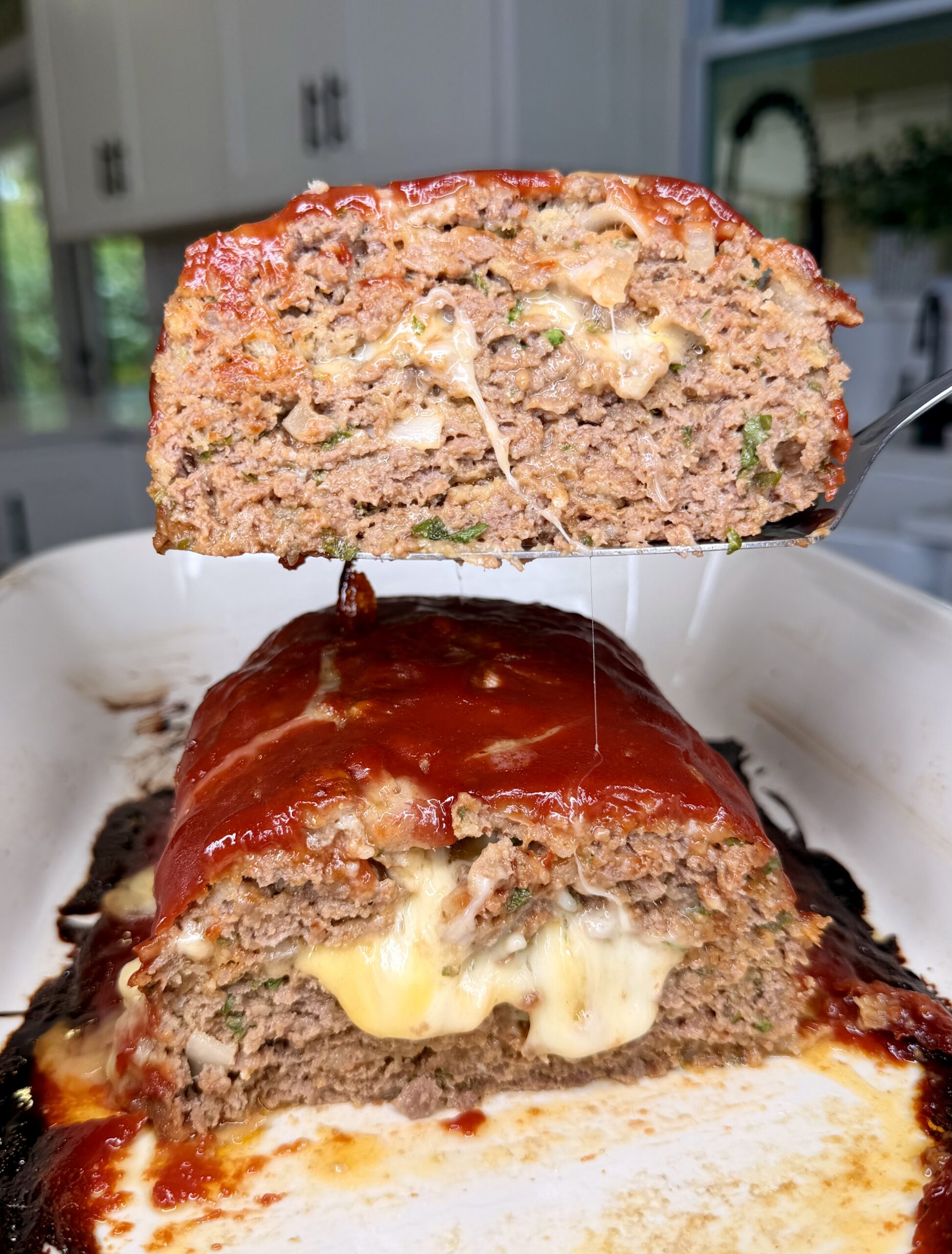 A sliced meatloaf in a white baking dish, topped with a glossy red sauce and filled with melted cheese, with one thick slice being lifted to show the cheesy center. A kitchen is visible in the background.
