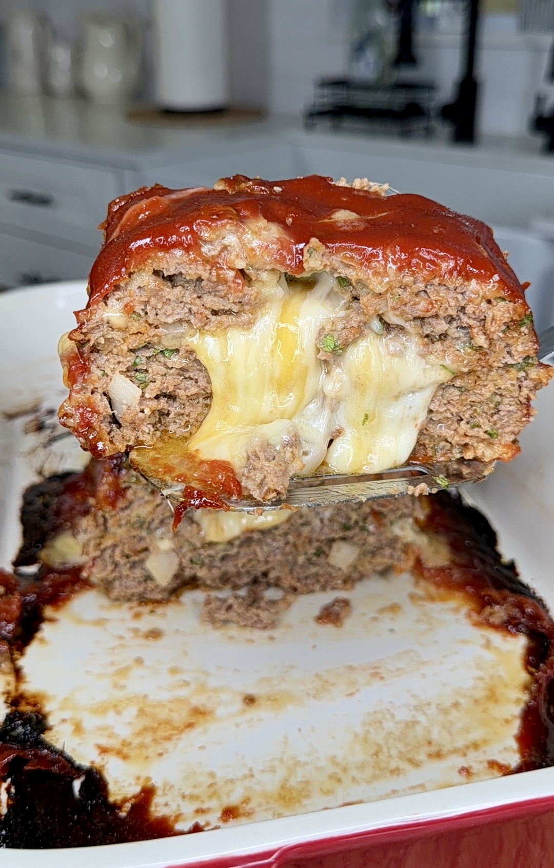 A slice of meatloaf filled with melted cheese being lifted from a baking dish, showing gooey cheese oozing out from the center. The meatloaf is topped with a glossy red sauce.