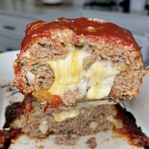 A close-up of a sliced meatloaf filled with melted cheese, topped with a glossy red glaze, with cheese oozing out from the center. The meatloaf sits on a white dish in a kitchen setting.