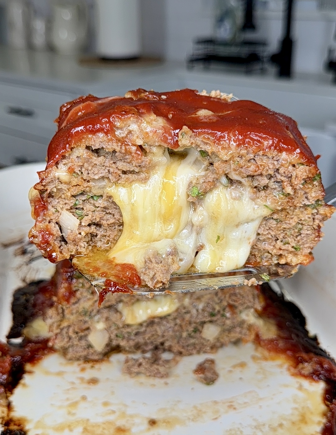 A close-up of a sliced meatloaf filled with melted cheese, topped with a glossy red glaze, with cheese oozing out from the center. The meatloaf sits on a white dish in a kitchen setting.