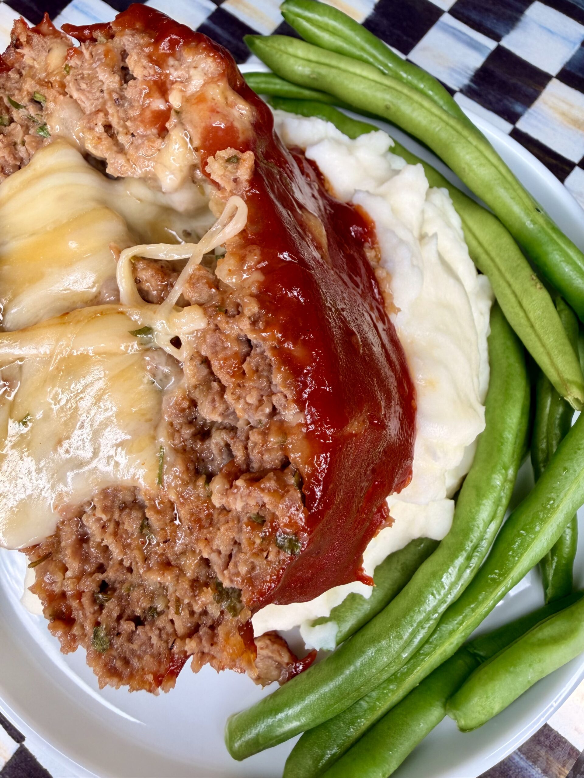 A close-up of a cheesy slice of meatloaf topped with tomato glaze, served on mashed potatoes with a side of fresh green beans on a white plate.