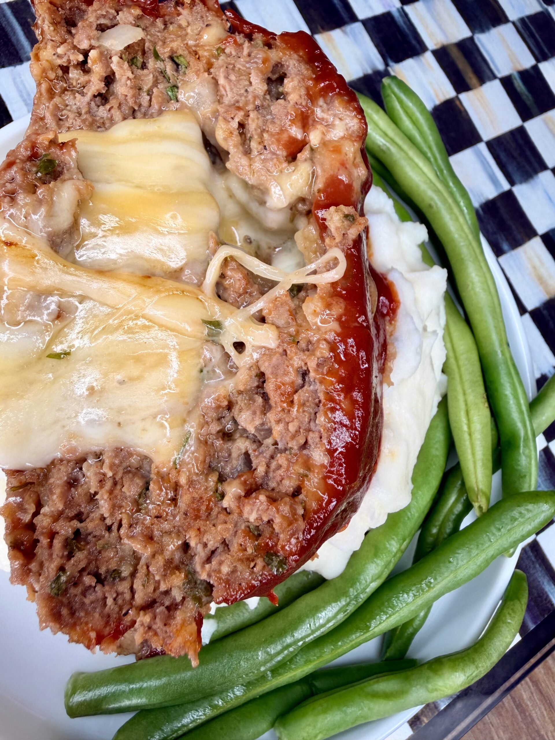 A slice of cheesy meatloaf topped with melted cheese sits on mashed potatoes, served with a side of green beans on a white plate with a checkered background.