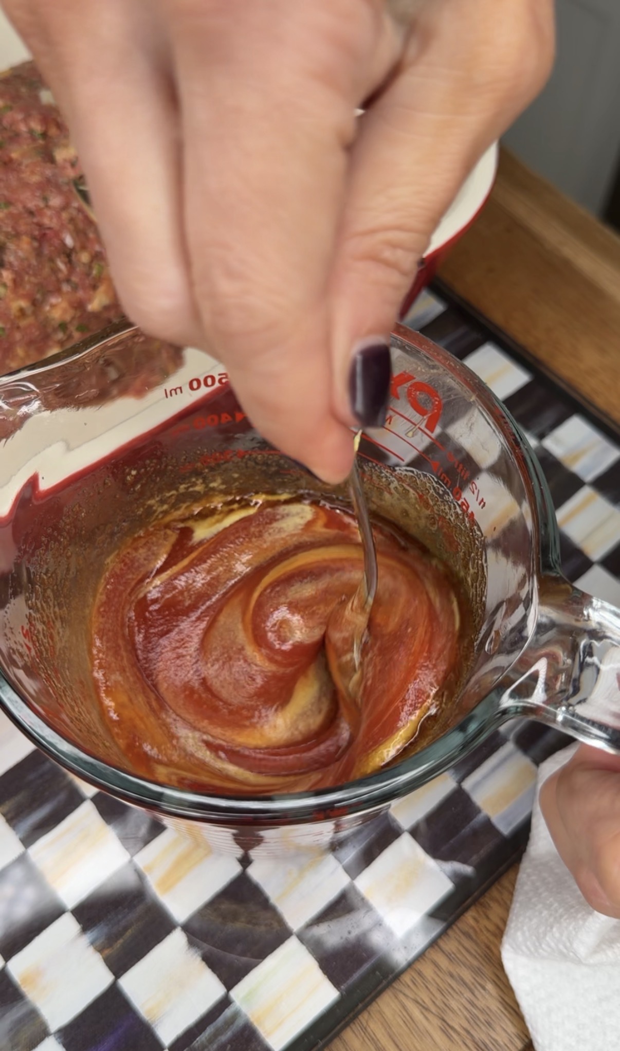 A hand stirs a reddish-brown sauce with a spoon in a glass measuring cup, blending the ingredients together. The cup is on a striped surface, and part of a meat mixture is visible in the background.