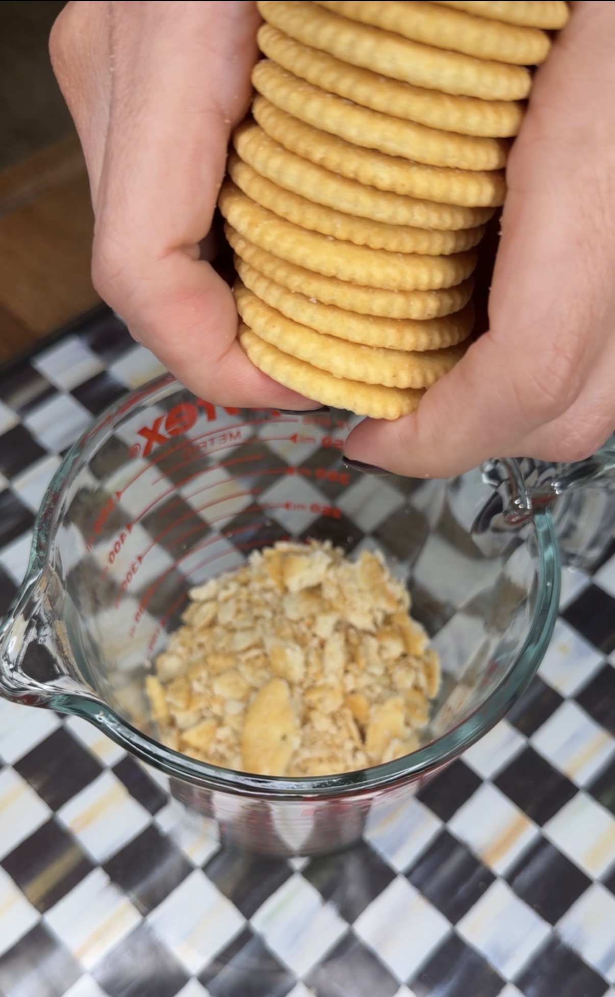 Hands hold a stack of round crackers above a glass measuring cup, which contains crushed crackers, on a checkered surface.