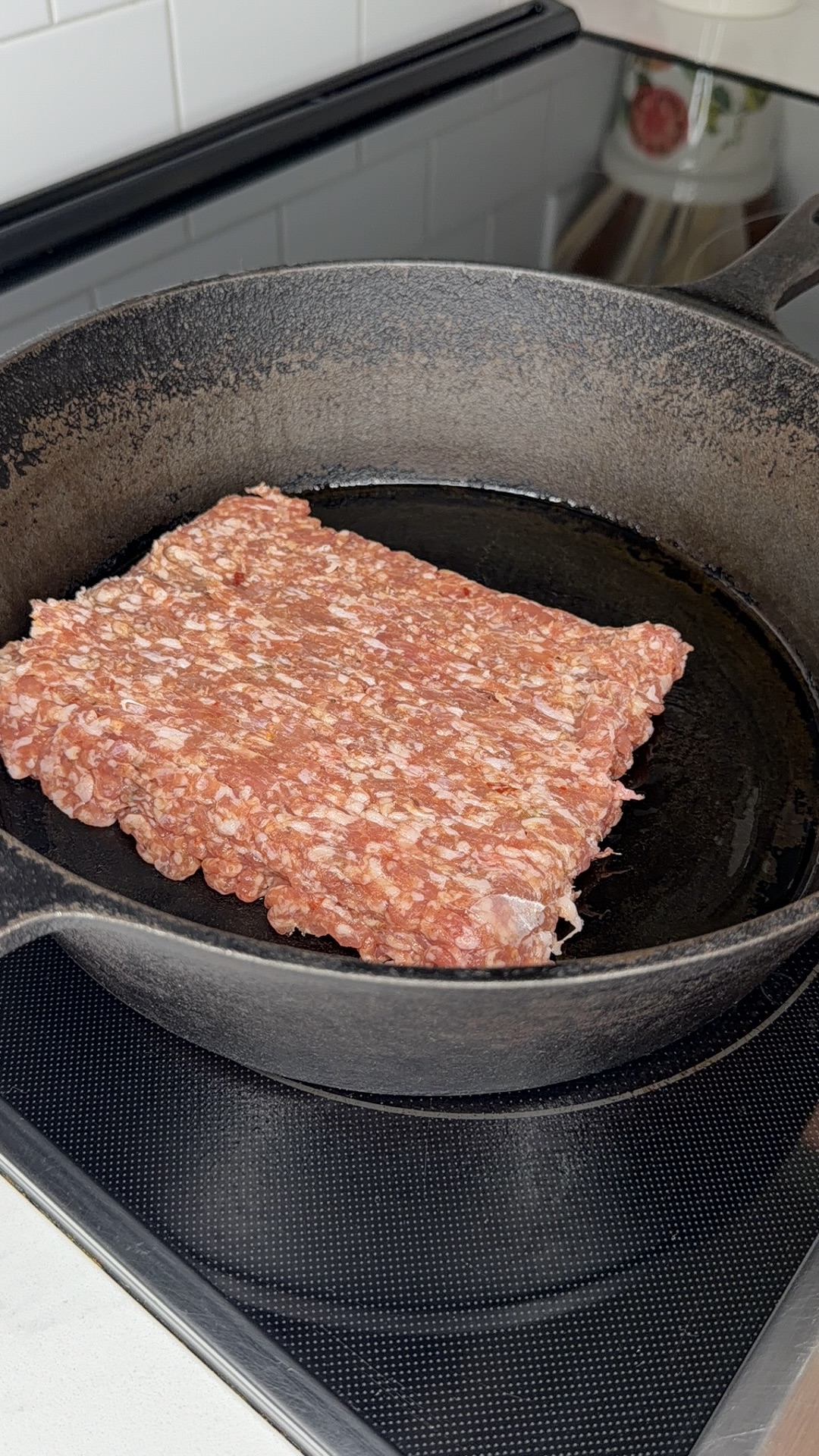 A rectangular block of raw ground meat is placed in a black cast iron skillet on a stovetop, ready for cooking.