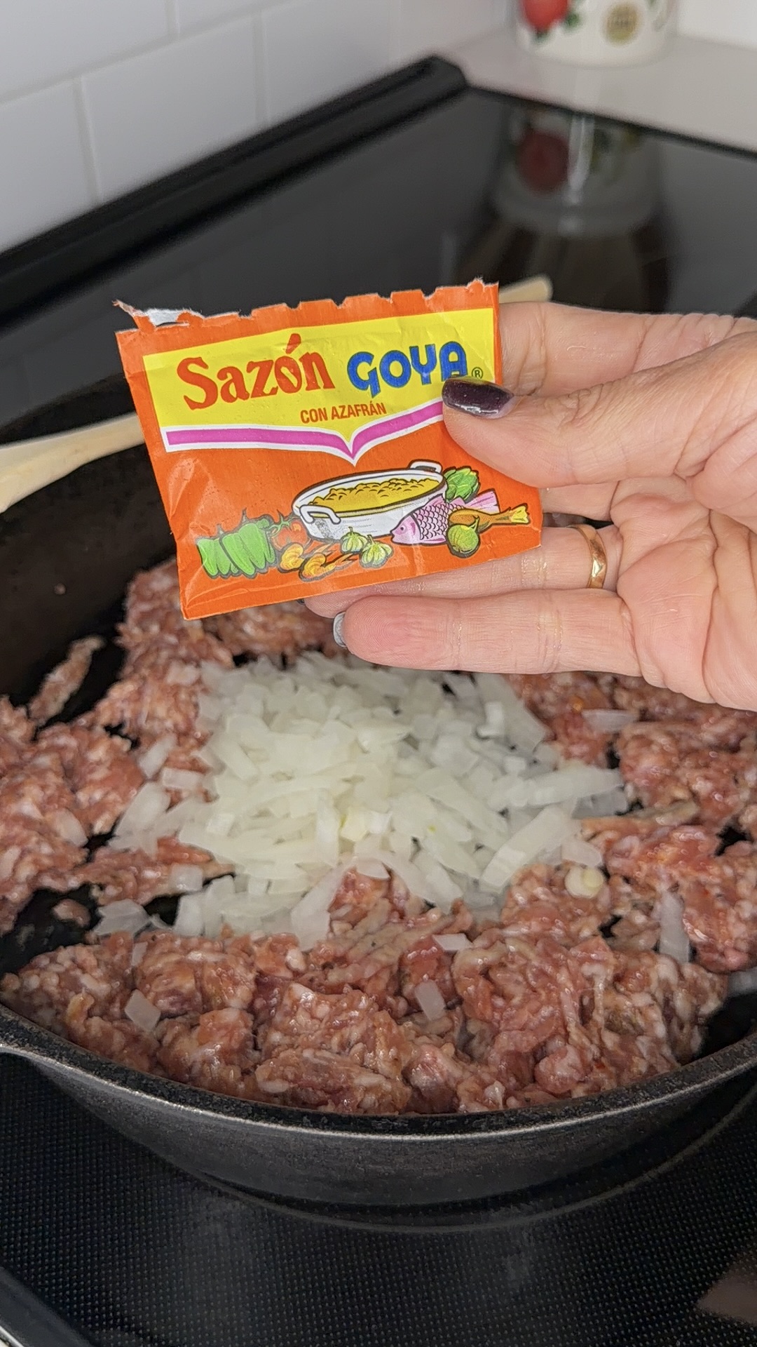 A hand holds a packet of Sazón Goya seasoning over a skillet with raw ground meat and chopped onions, ready to be cooked, with a wooden spoon resting on the pan.