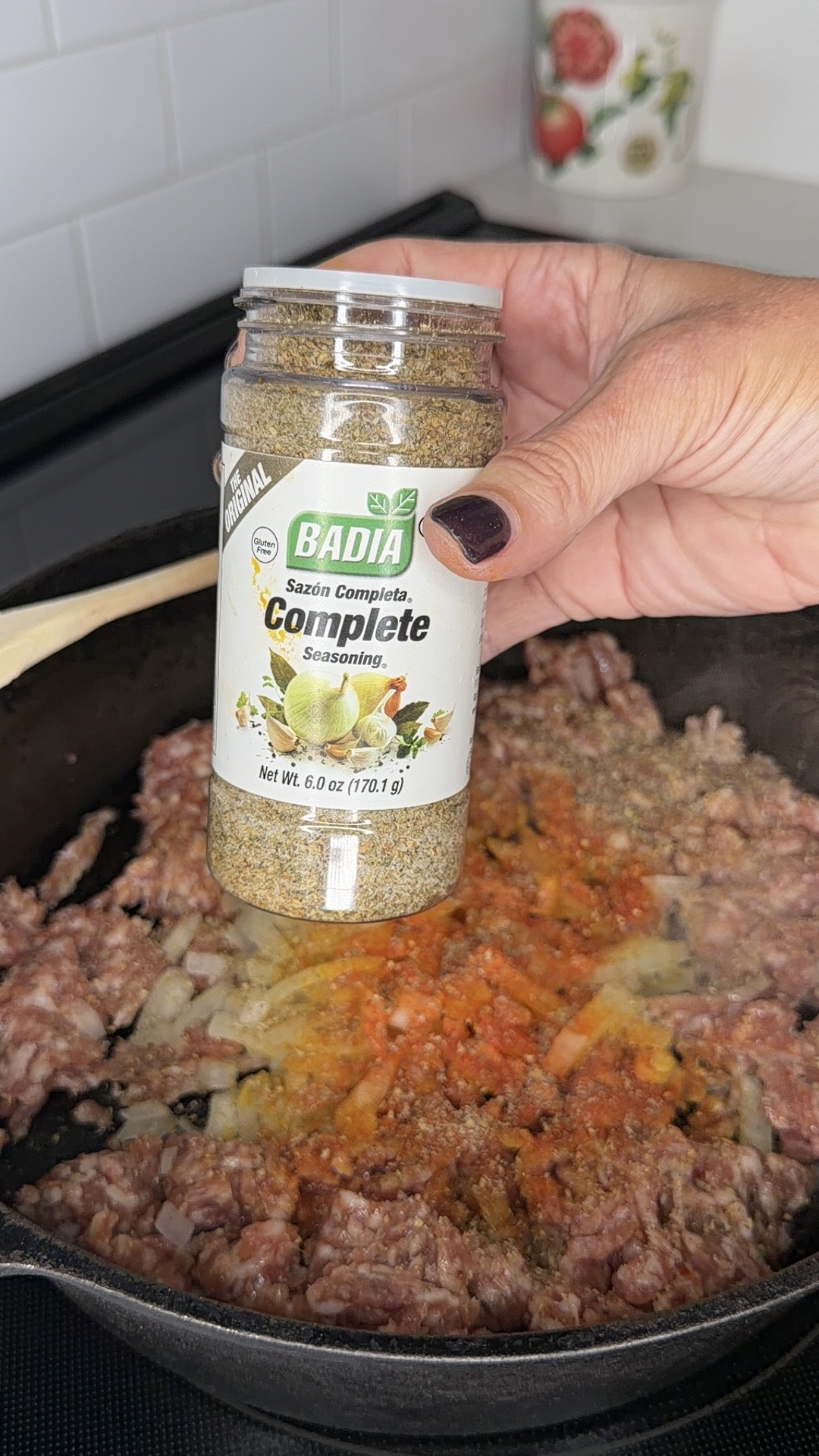 A hand sprinkles Badia Complete Seasoning over ground meat and diced onions cooking in a black skillet on a stovetop.