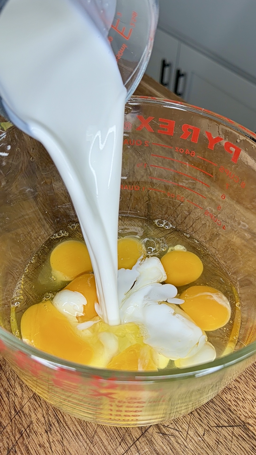 Milk is being poured from a measuring cup into a glass bowl containing several cracked eggs, sitting on a wooden surface. The bowl is labeled “Pyrex” and kitchen cabinets are visible in the background.