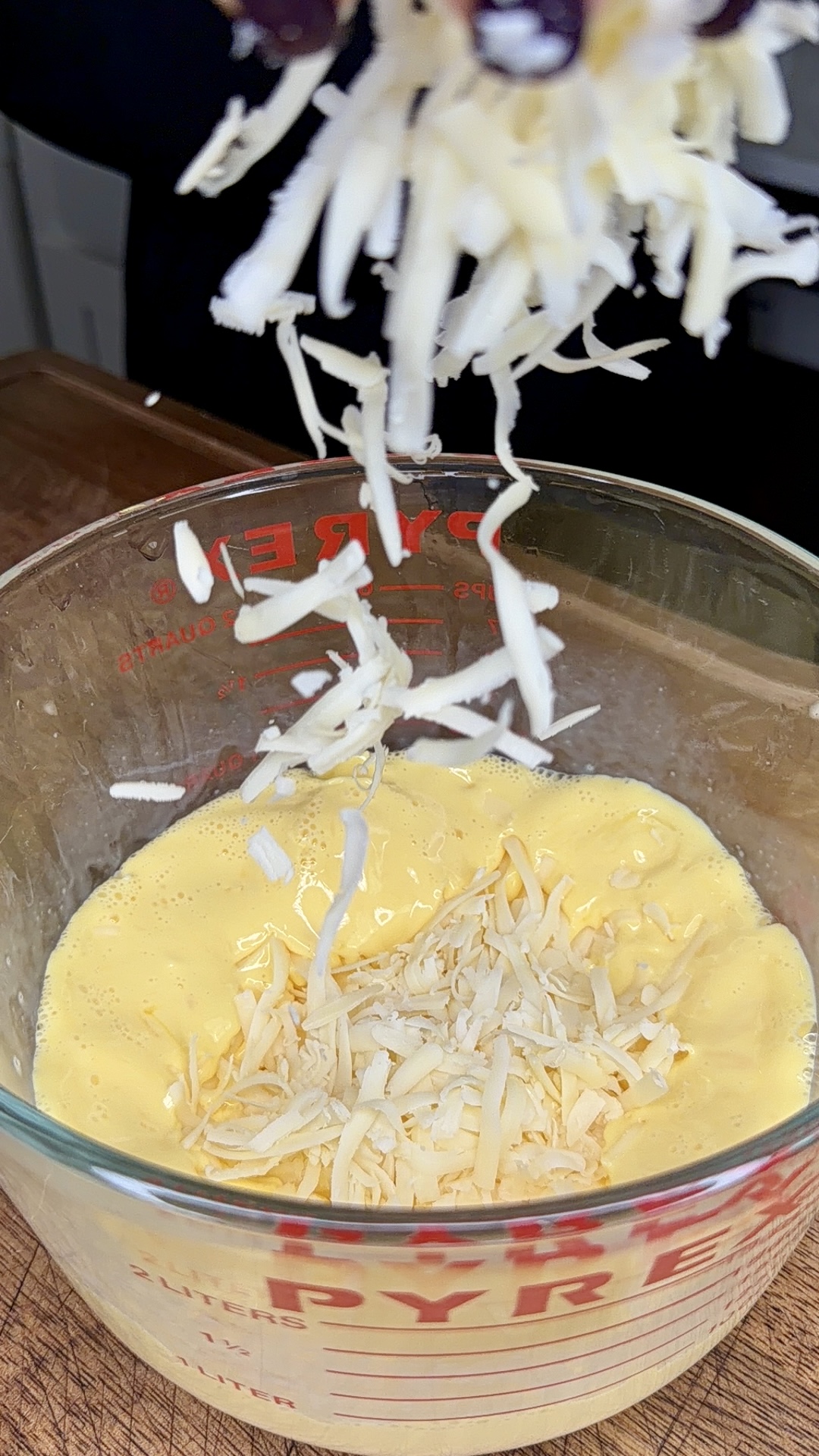 Shredded cheese is being sprinkled into a glass measuring cup filled with beaten eggs, sitting on a wooden surface.