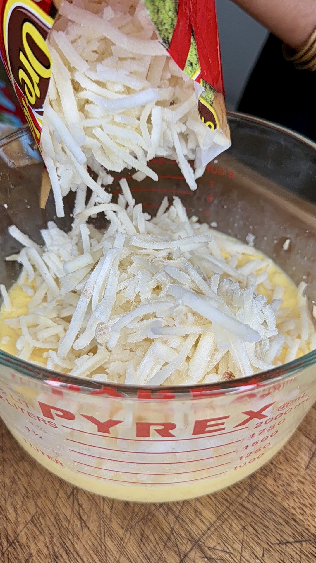 A bag of shredded potatoes is being poured into a glass Pyrex measuring cup containing beaten eggs, on a wooden surface.