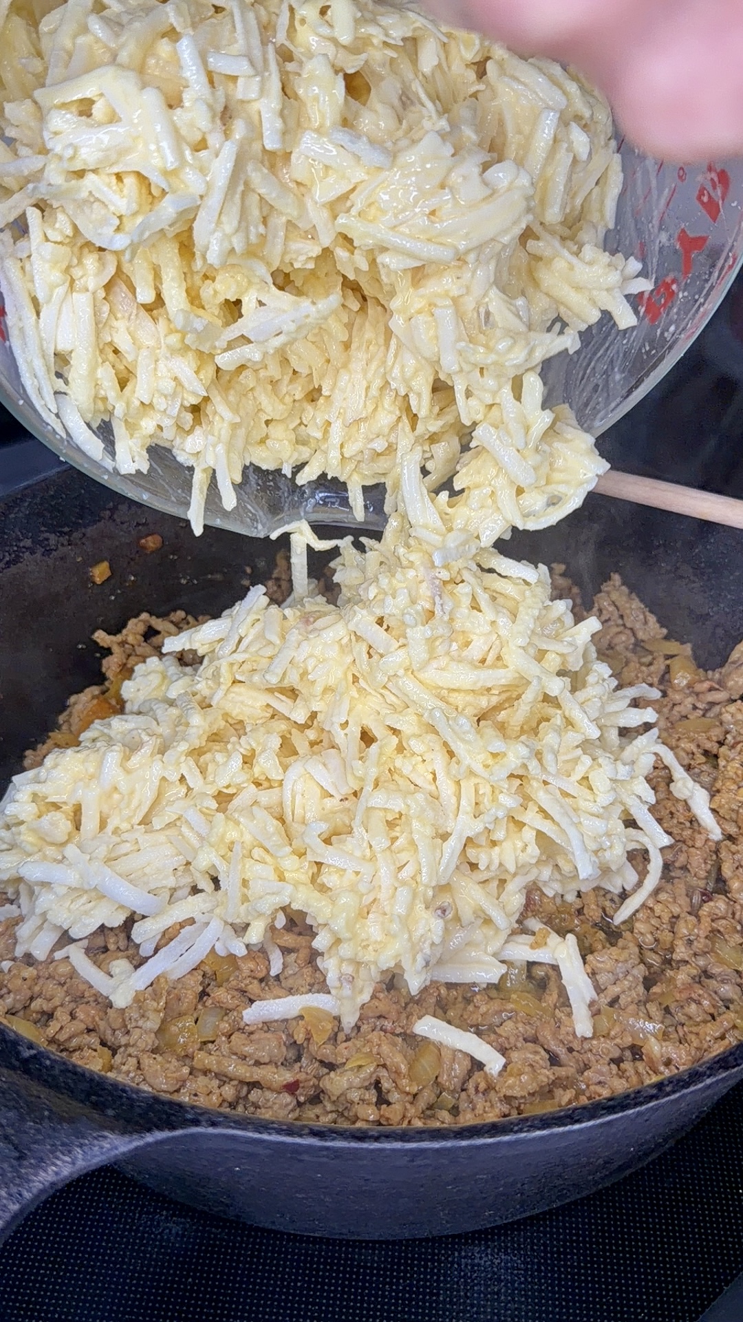 A bowl of shredded hash browns mixed with cheese is being poured into a cast iron skillet filled with cooked ground beef.