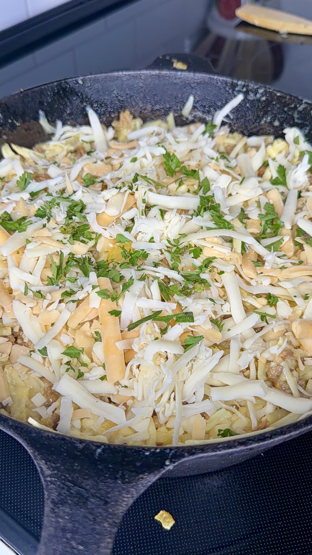 A close-up of a cast iron skillet filled with a dish topped with shredded cheese and chopped fresh herbs, ready to be cooked or baked.