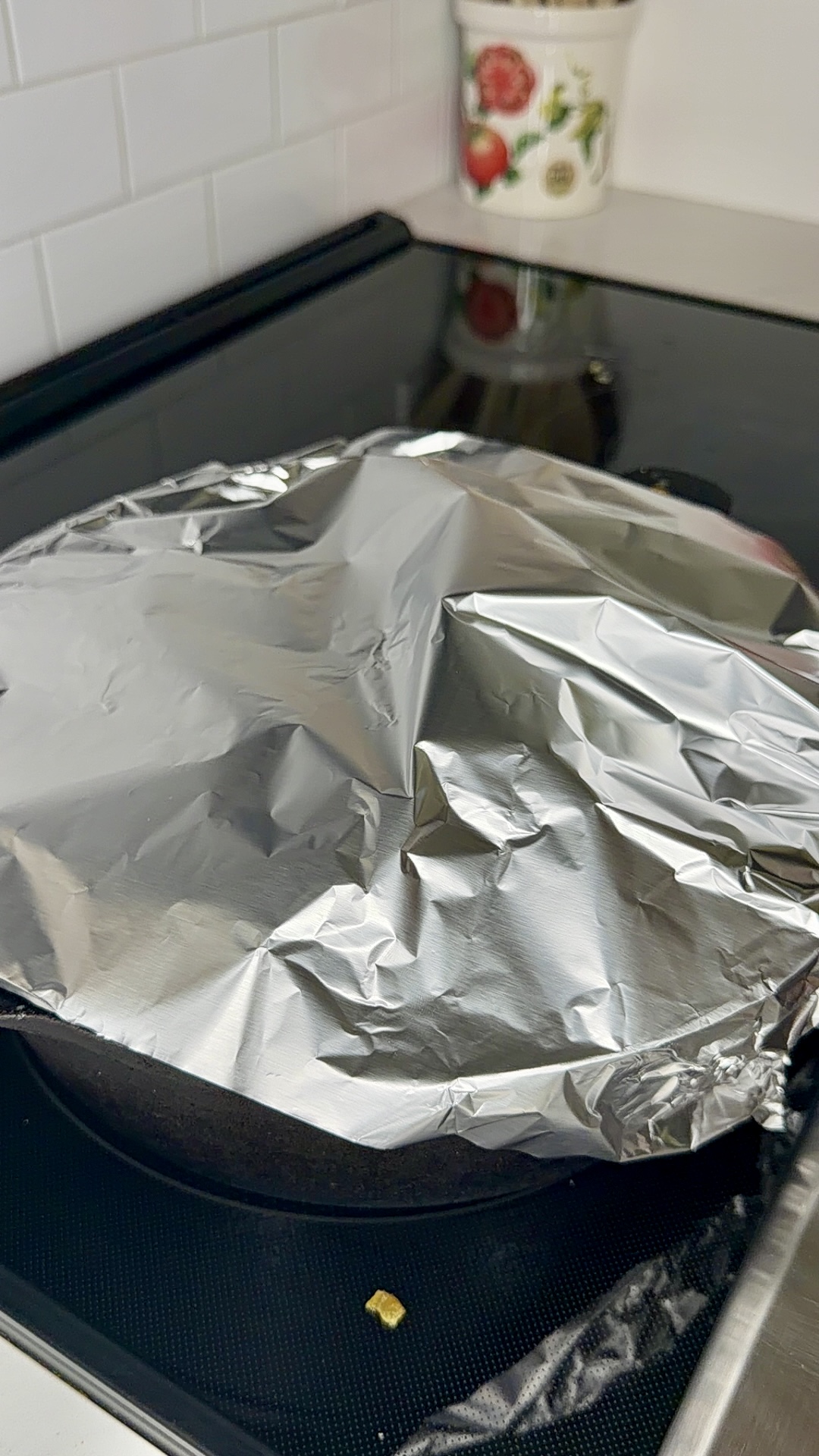 A stovetop pan covered with crumpled aluminum foil sits on a black glass cooktop. In the background, a floral-patterned container is on the white countertop against a white tile wall.