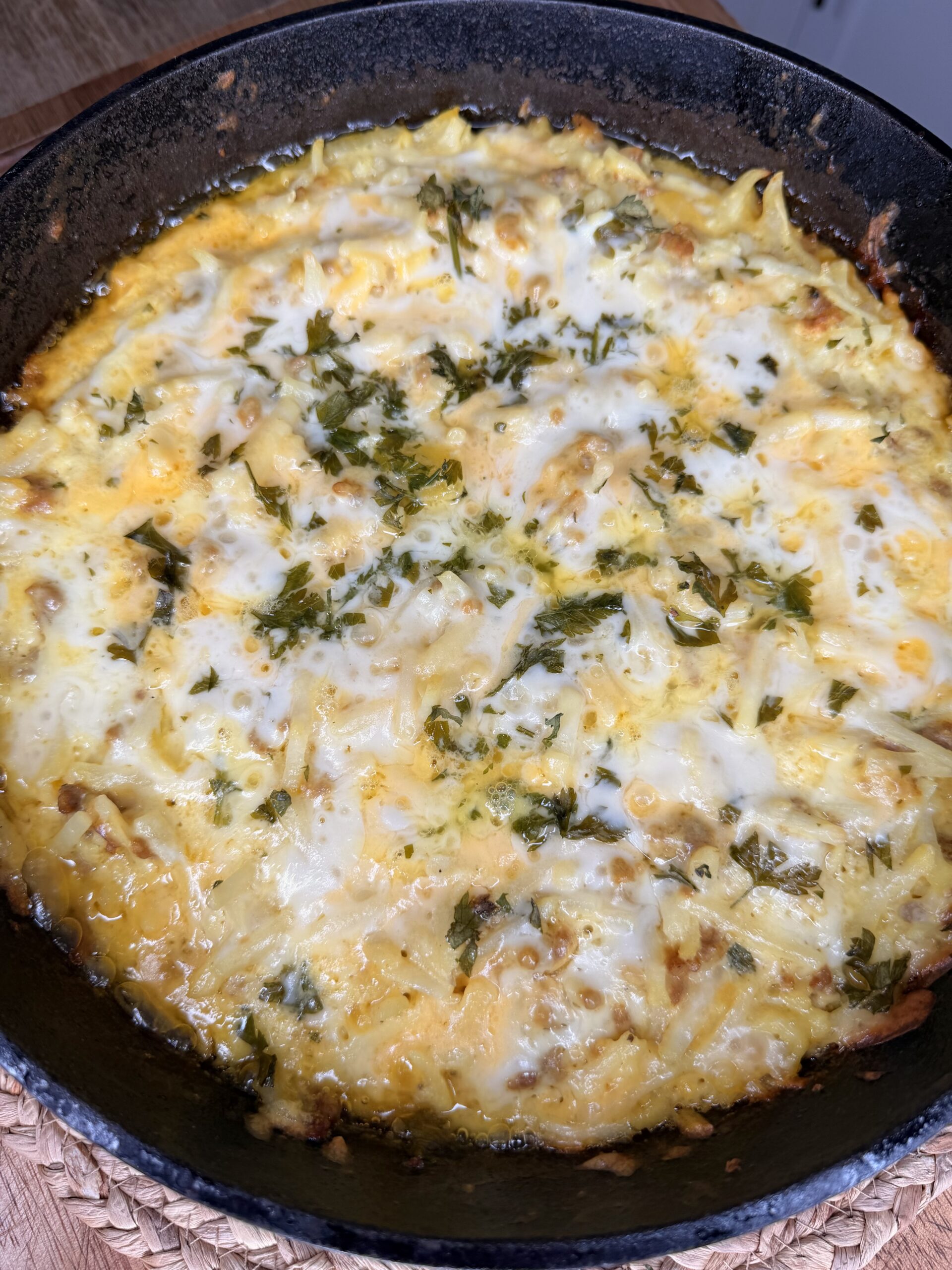 A cast iron skillet filled with a baked cheesy casserole, topped with melted cheese and sprinkled with chopped fresh herbs.