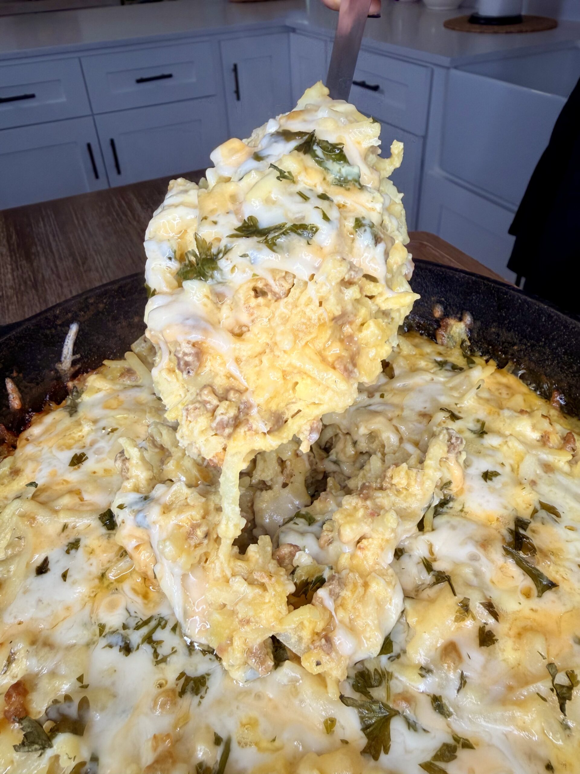 A close-up of a cheesy casserole being served from a skillet, showing melted cheese, herbs, and a creamy, golden texture. The kitchen in the background has white cabinets and wooden countertops.