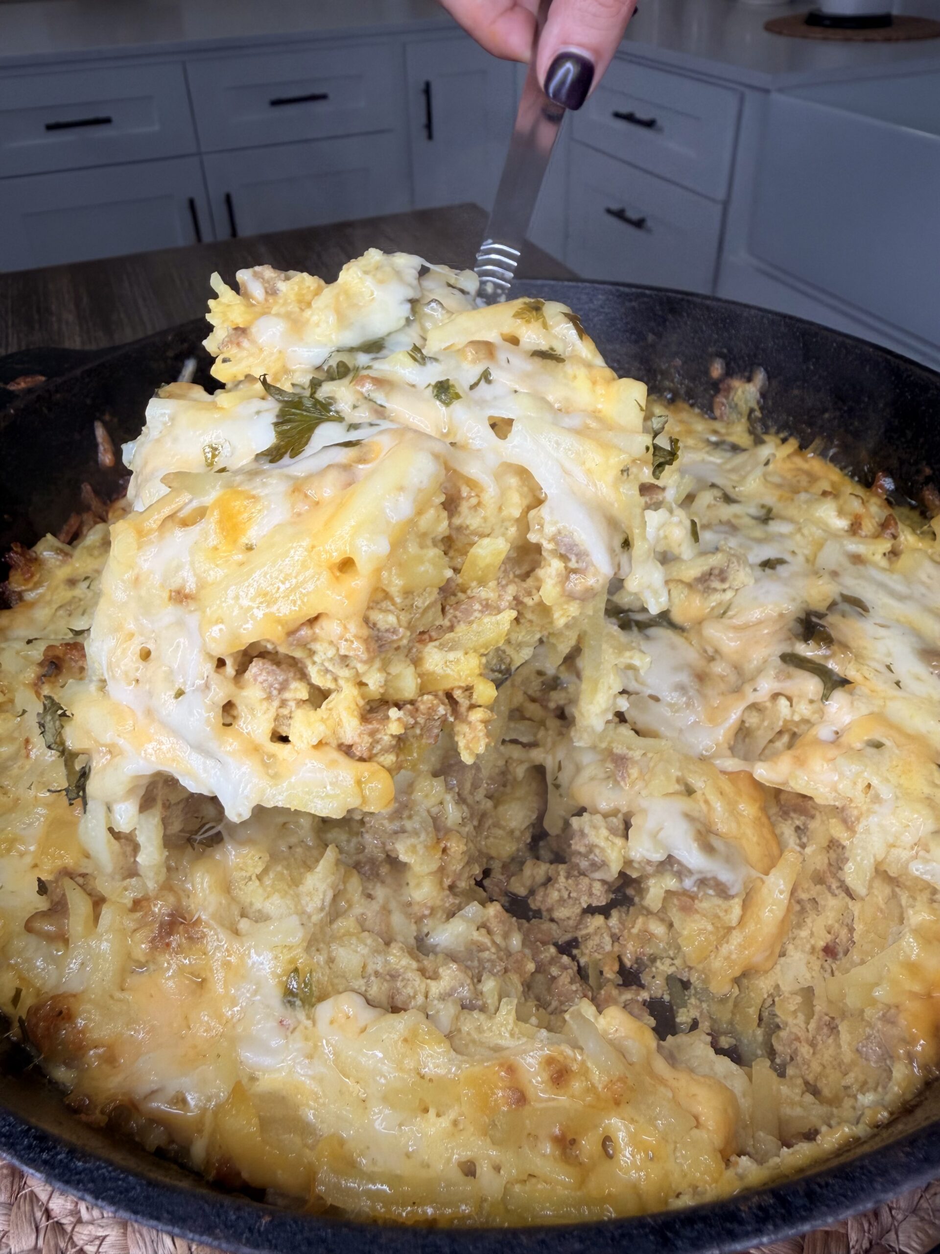 A close-up of a cheesy, golden-baked casserole with ground meat and herbs being served from a cast iron skillet in a kitchen. A hand holds a spatula lifting a large portion from the pan.