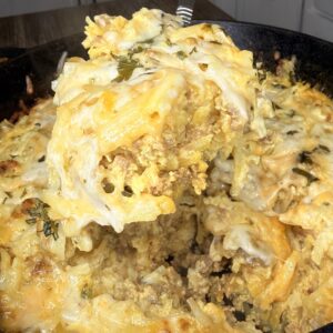 A close-up of a cheesy casserole being served from a cast iron skillet, showing layers of ground meat, melted cheese, potatoes, and herbs. The creamy, golden dish is set against a kitchen background.