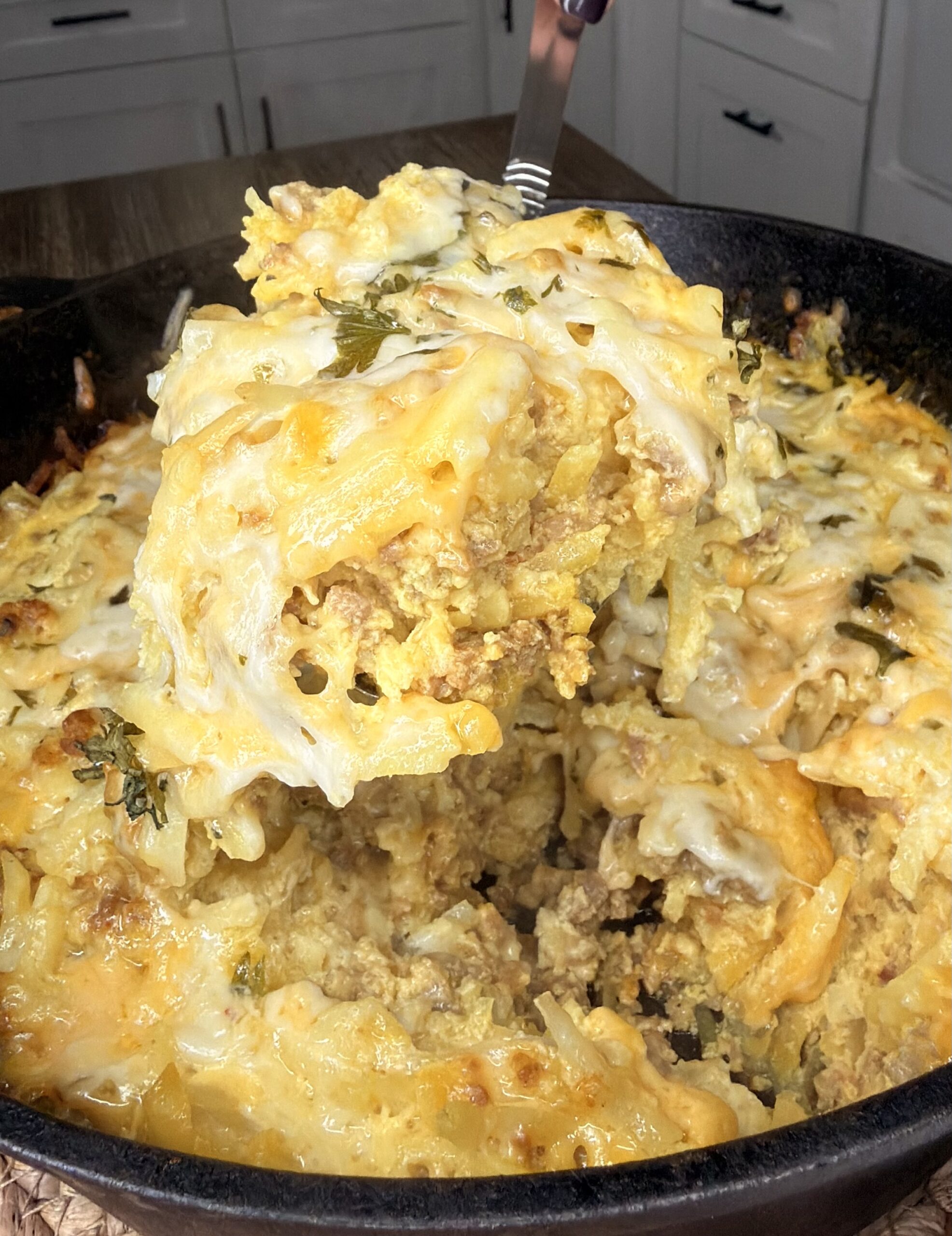 A close-up of a cheesy casserole being served from a cast iron skillet, showing layers of ground meat, melted cheese, potatoes, and herbs. The creamy, golden dish is set against a kitchen background.