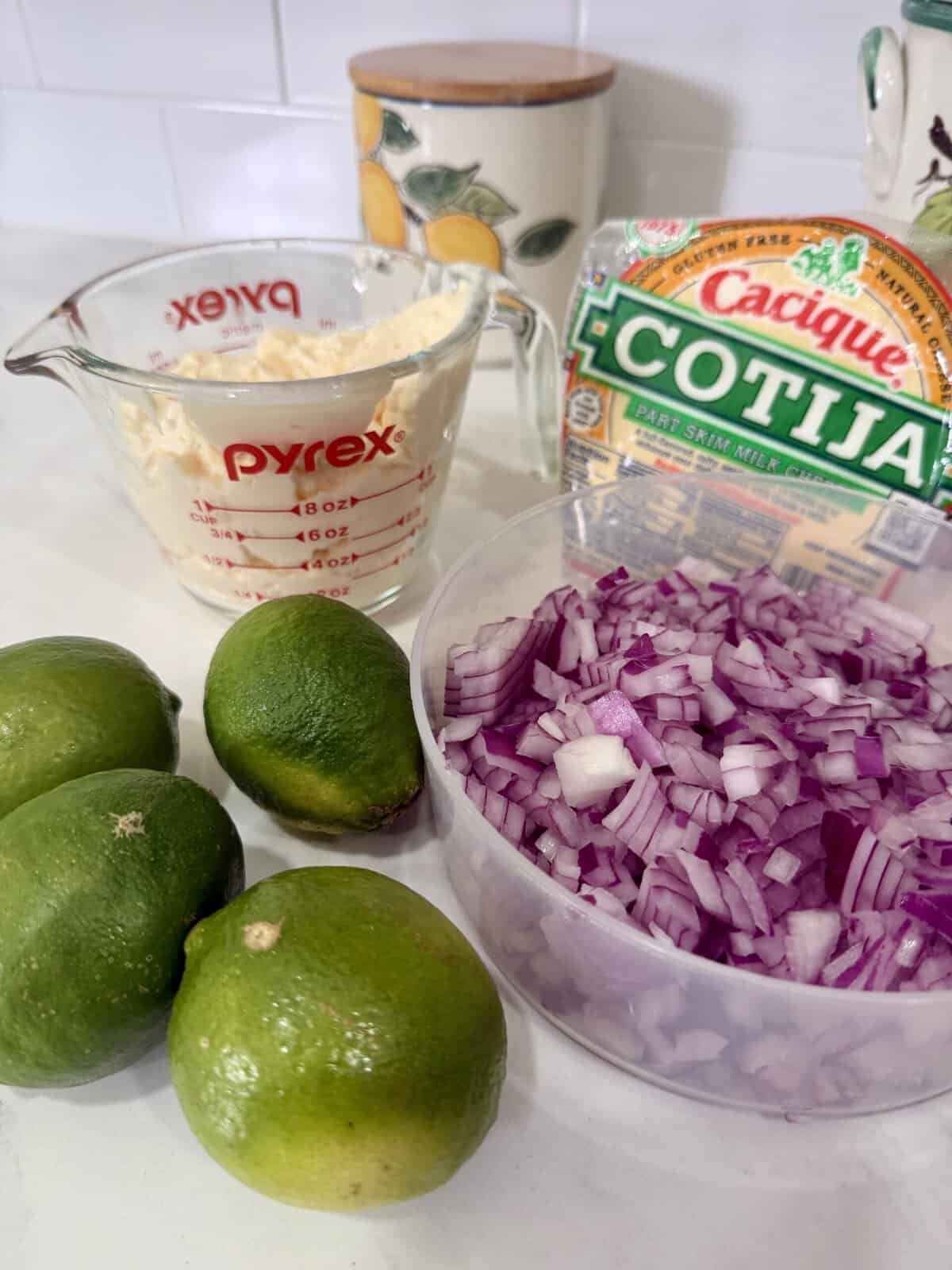 A kitchen counter with four limes, a container of chopped red onion, a measuring cup of mayonnaise, and a package of Cotija cheese. A decorative container with a lemon design is in the background.