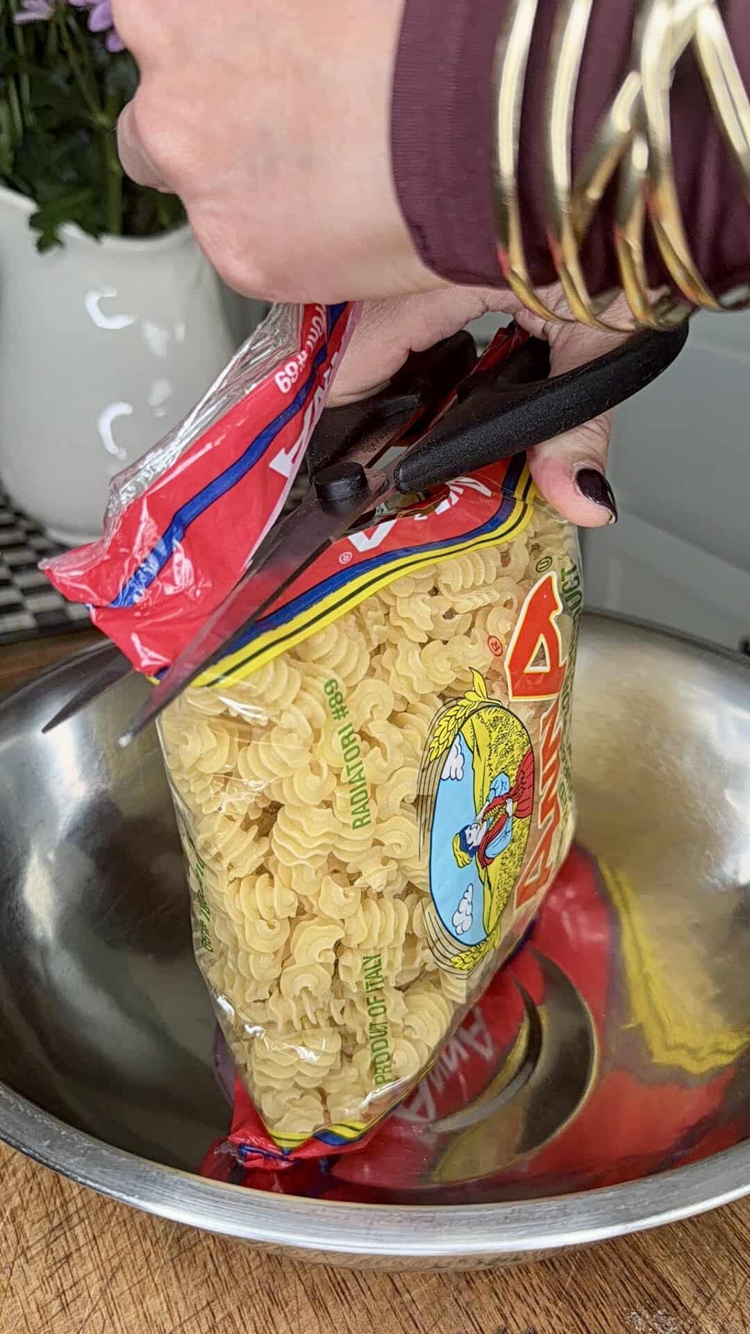 A hand holding scissors cuts open a bag of uncooked spiral pasta over a metal bowl. The arm wears gold bangles and has dark-colored nail polish.