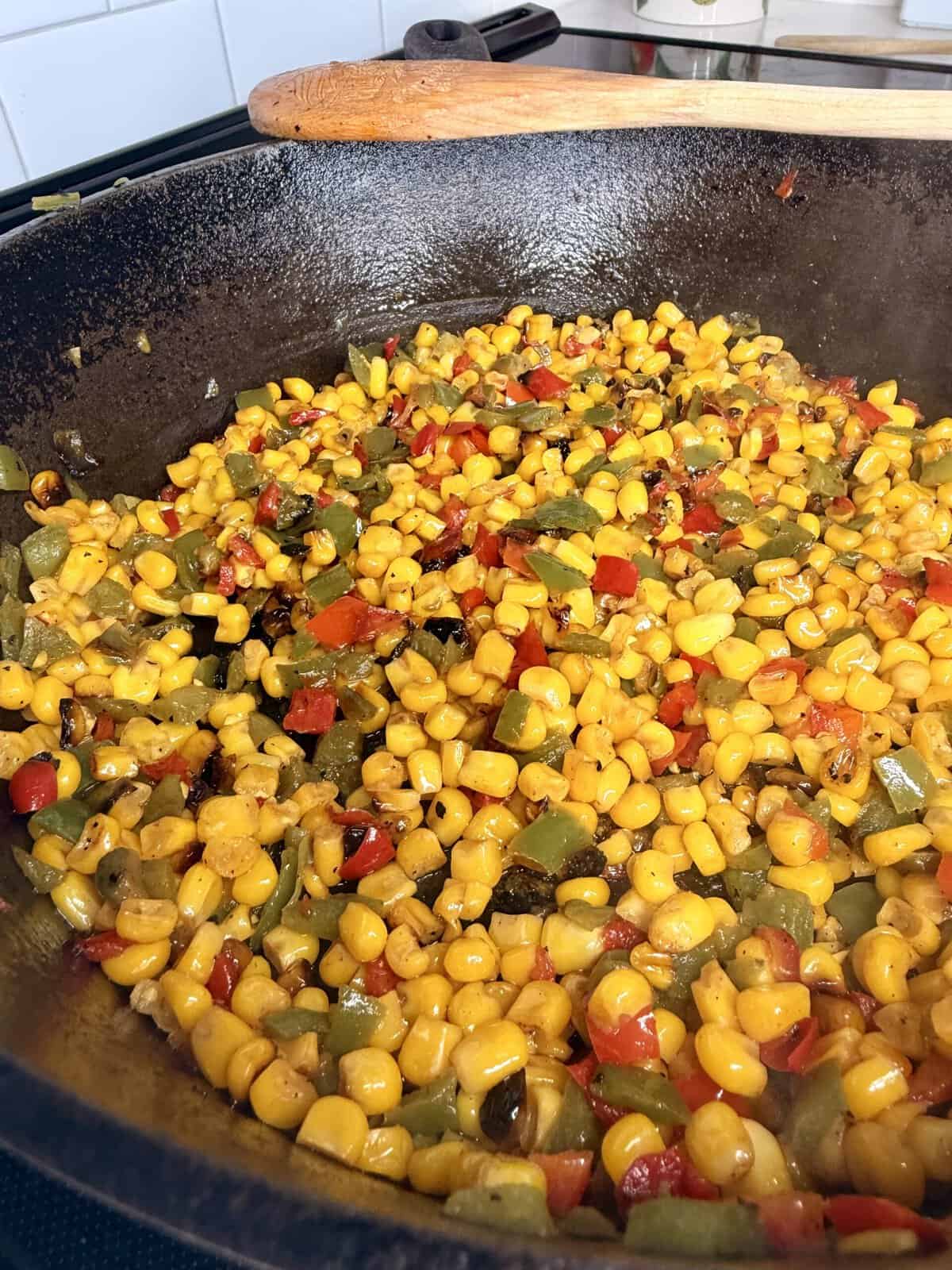 Colorful sautéed corn, red and green bell peppers, and onions cooking in a cast iron skillet on a stovetop, with a wooden spatula resting on the rim.