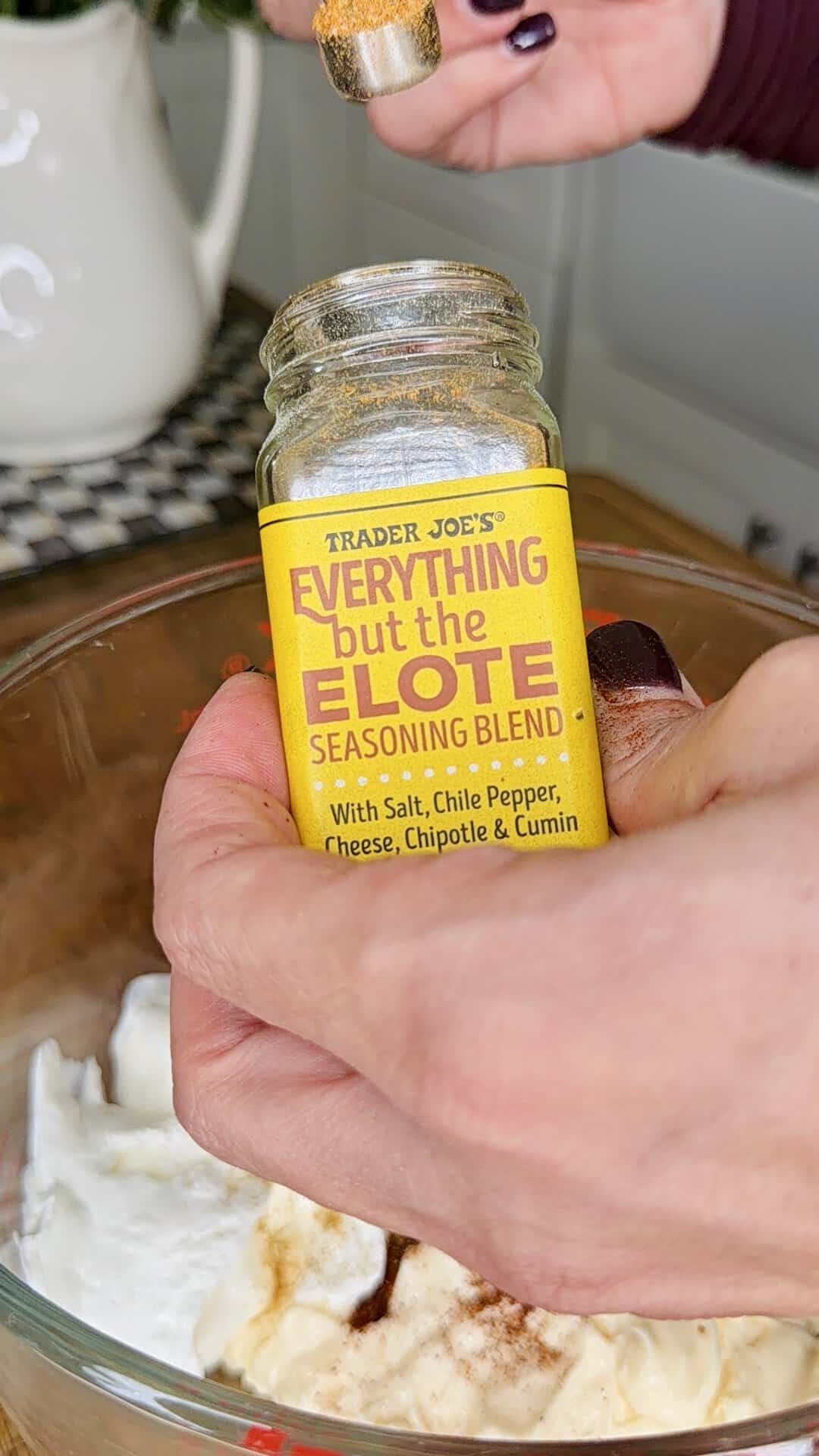 A hand sprinkles Trader Joe’s Everything but the Elote Seasoning Blend over a bowl of white creamy mixture in a kitchen setting.