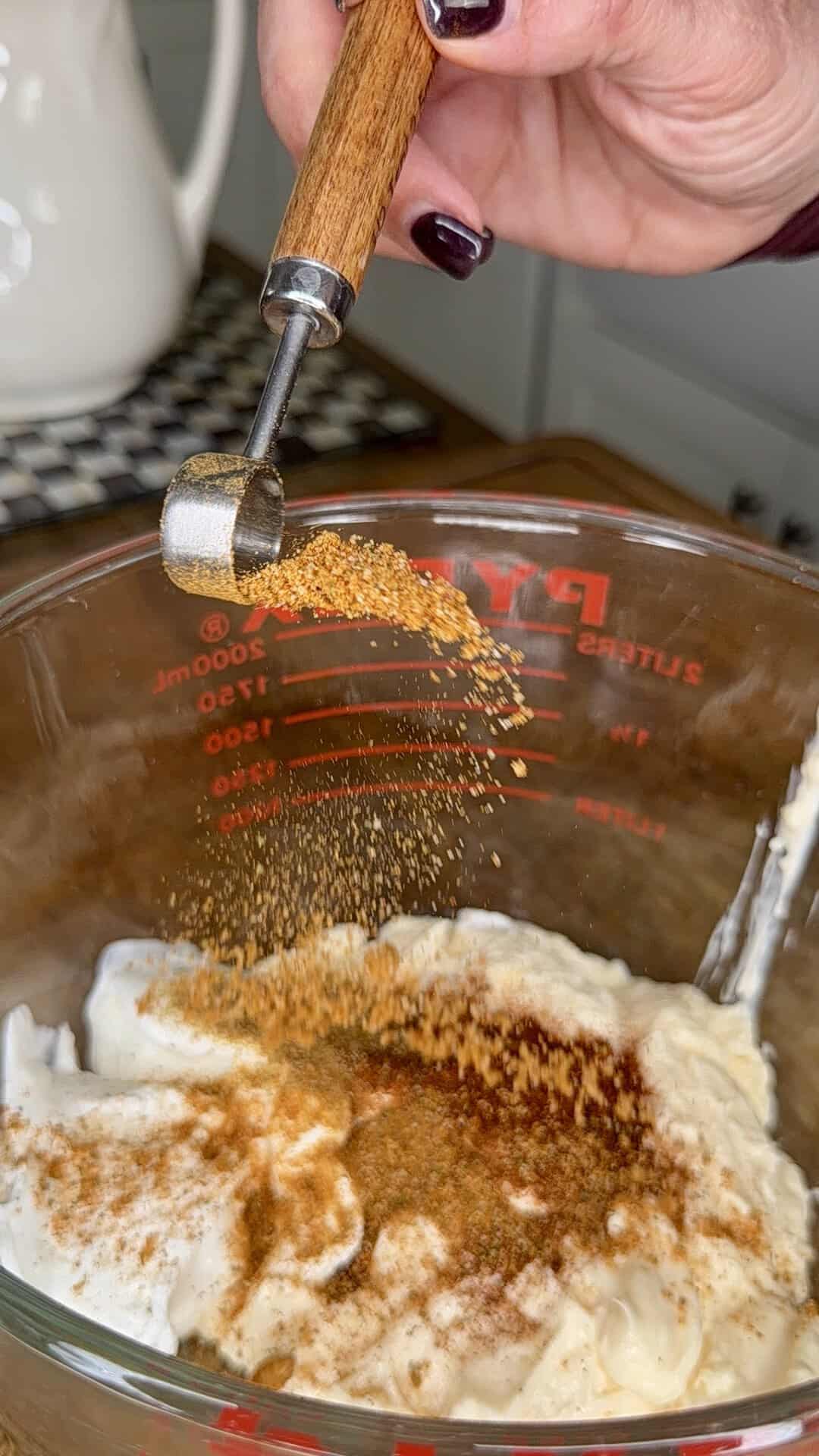A hand sprinkles seasoning from a small wooden-handled spoon into a glass measuring cup filled with a creamy mixture in a kitchen setting.