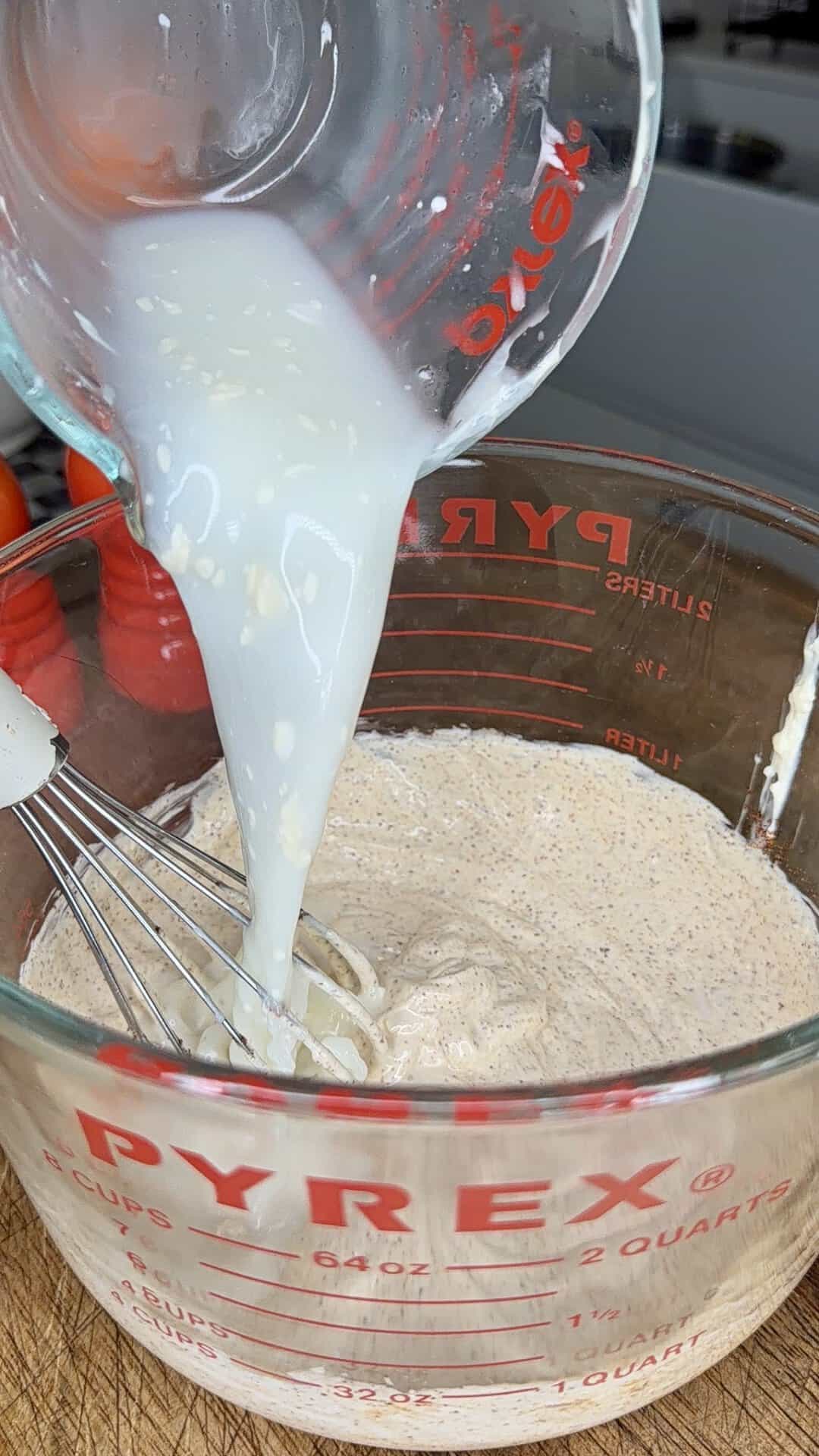 A glass measuring cup pours a white liquid into another glass measuring cup containing a beige, speckled mixture being whisked. Both cups are labeled Pyrex.