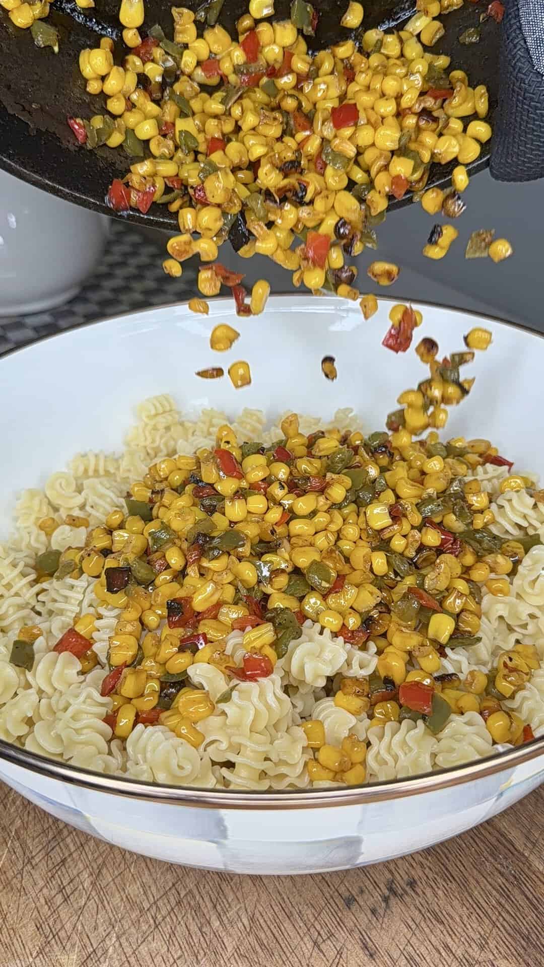 Charred corn and diced red and green peppers are being poured from a pan onto a bowl filled with cooked ruffle-edged pasta on a wooden surface.