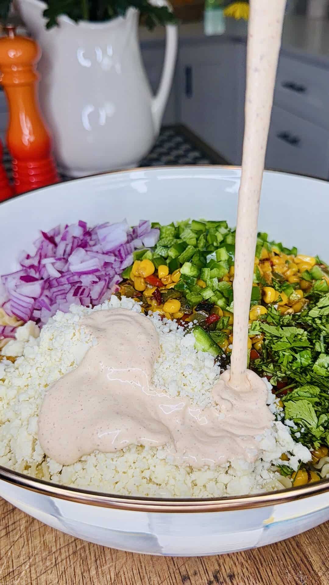 A bowl filled with chopped red onion, green peppers, corn, herbs, crumbled cheese, and a creamy sauce being poured on top, set on a wooden surface in a kitchen.