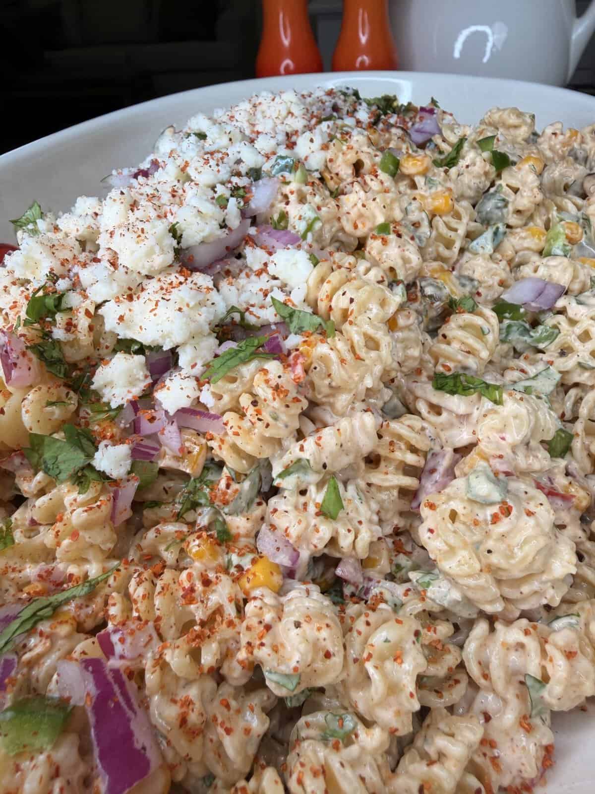 A close-up of a creamy pasta salad with rotini, chopped red onions, cilantro, crumbled cheese, corn, and a sprinkle of chili powder, served in a white dish with salt and pepper shakers in the background.