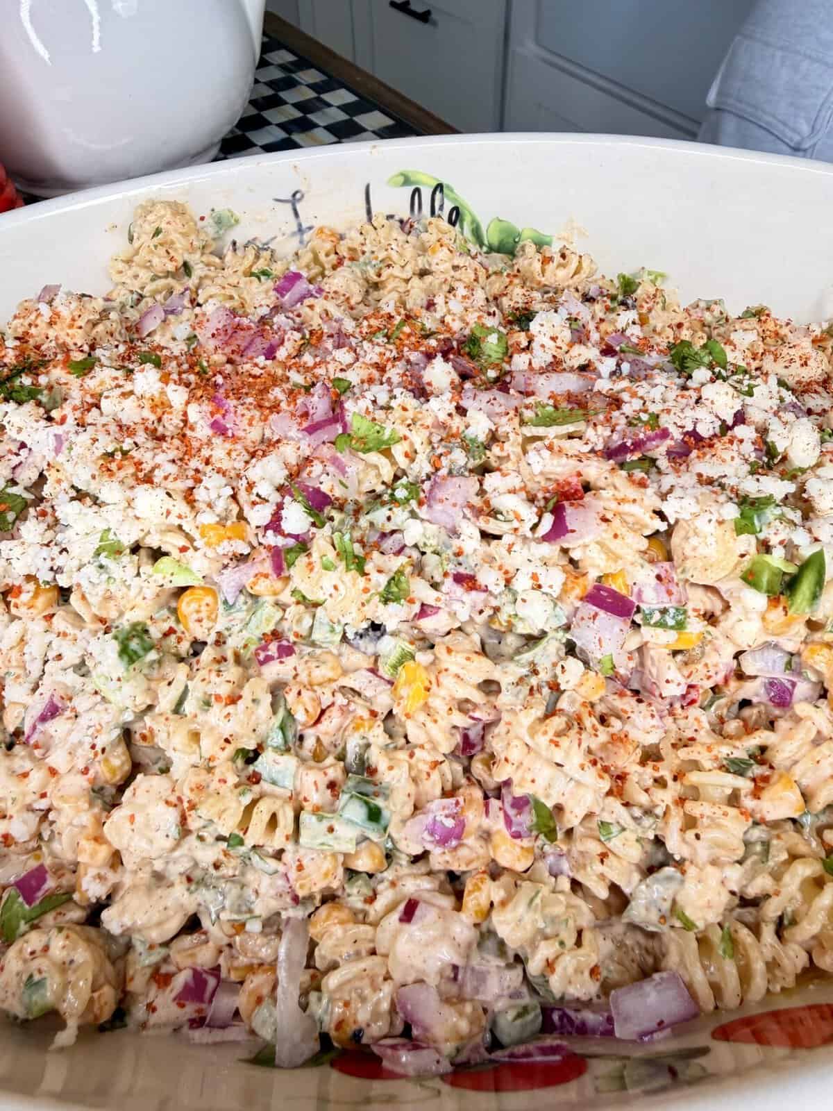 A close-up of a large bowl of pasta salad with rotini noodles, chopped red onions, green peppers, corn, crumbly cheese, and a sprinkle of paprika on top. The salad appears creamy and colorful.