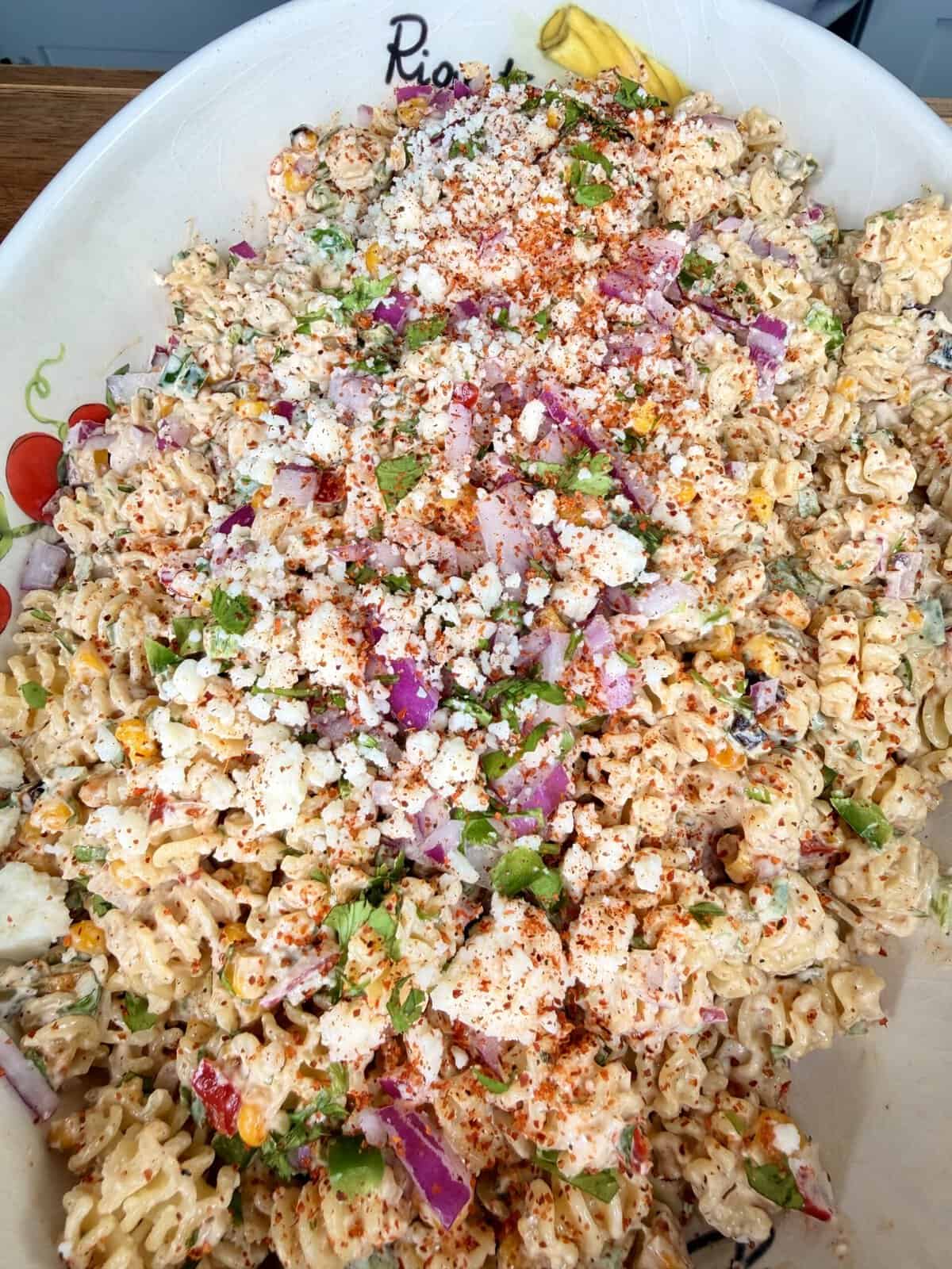 A large bowl of rotini pasta salad mixed with crumbled cheese, chopped red onions, green herbs, and sprinkled with red chili powder or paprika. The salad appears creamy and colorful.