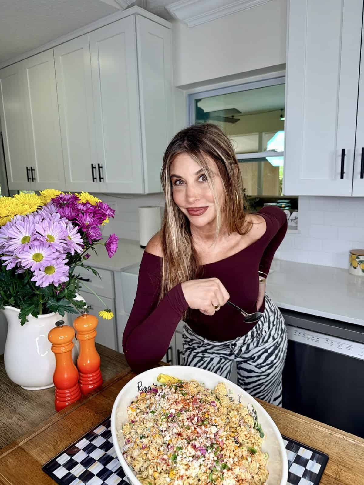 A woman with long brown hair in an off-shoulder top smiles while holding a fork over a large bowl of colorful salad on a kitchen counter, with flowers and orange salt and pepper shakers beside her.