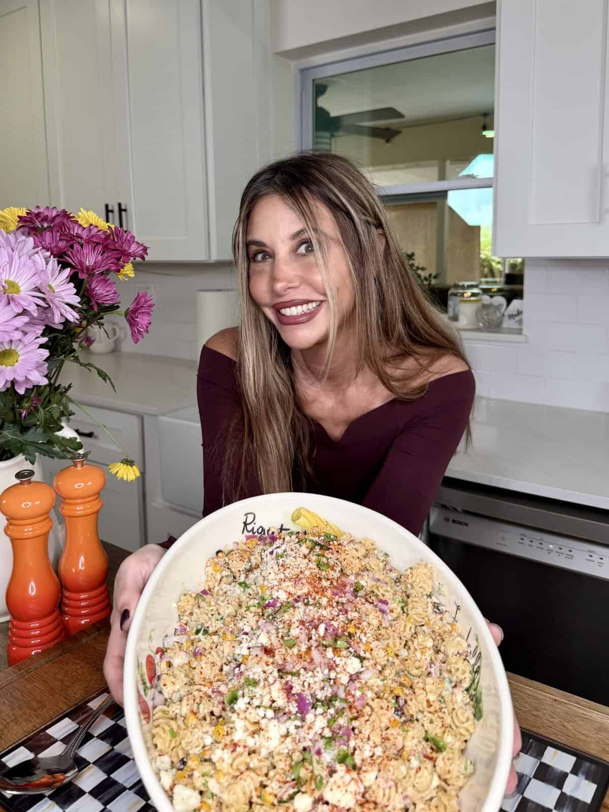 A woman with long brown hair, wearing a maroon top, smiles while holding a large bowl of pasta salad topped with herbs and cheese in a bright kitchen with white cabinets and a vase of flowers.