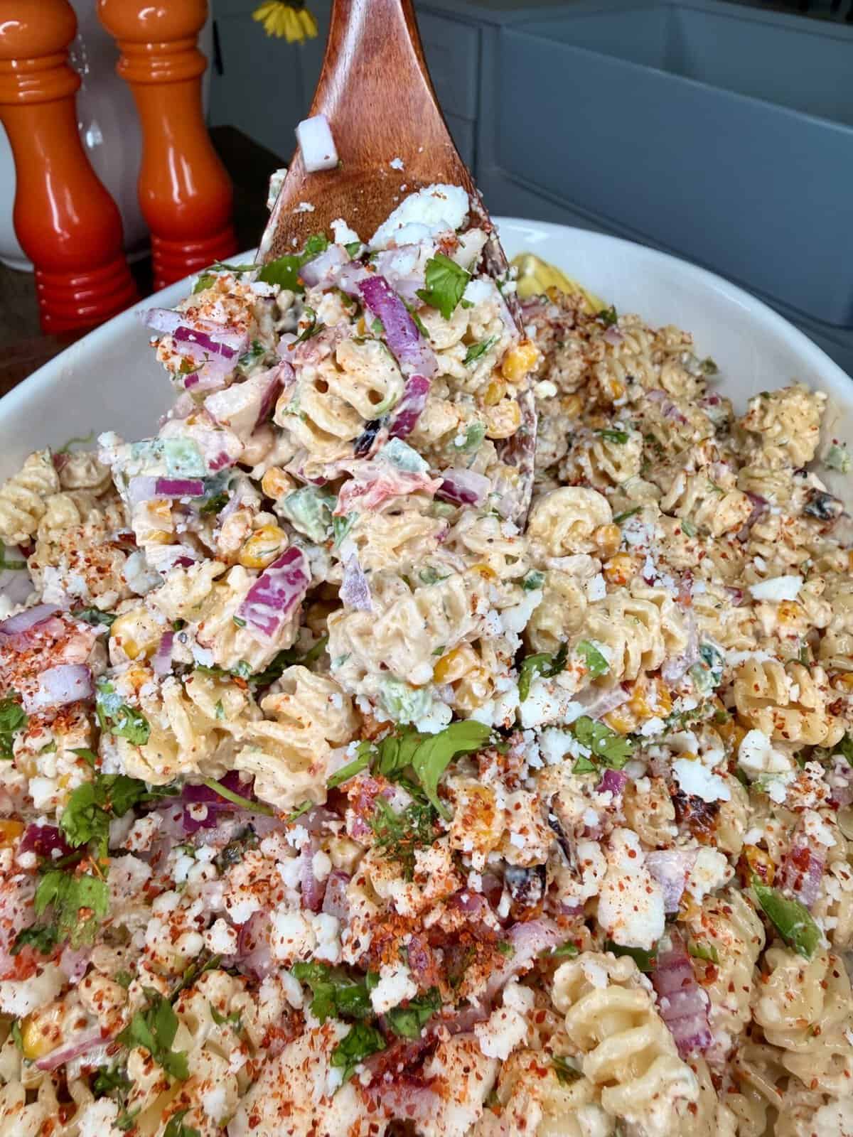 A wooden spoon scoops up creamy pasta salad with rotini noodles, red onion, corn, cilantro, cheese, and sprinkled spices from a white dish. Two pepper grinders are visible in the background.