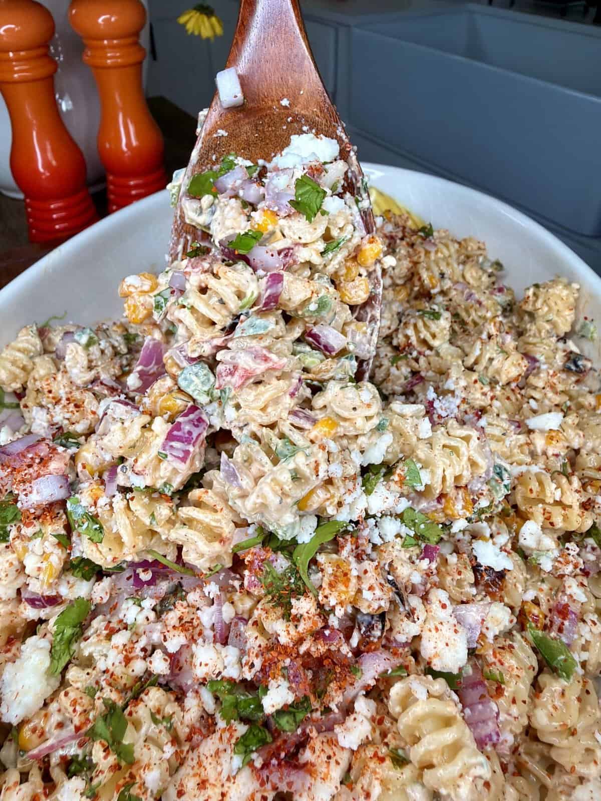 A serving spoon lifts a portion of creamy pasta salad with rotini, red onions, corn, cilantro, cheese, and sprinkled spices from a large white dish. Red and orange pepper grinders are visible in the background.