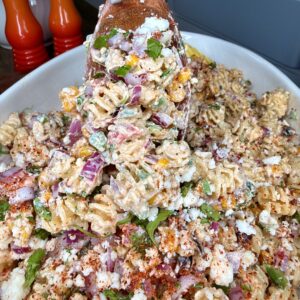 A wooden spoon lifts a serving of creamy pasta salad with rotini, red onions, green peppers, cilantro, crumbled cheese, and sprinkled spices from a large white dish. Two orange pepper mills are visible in the background.