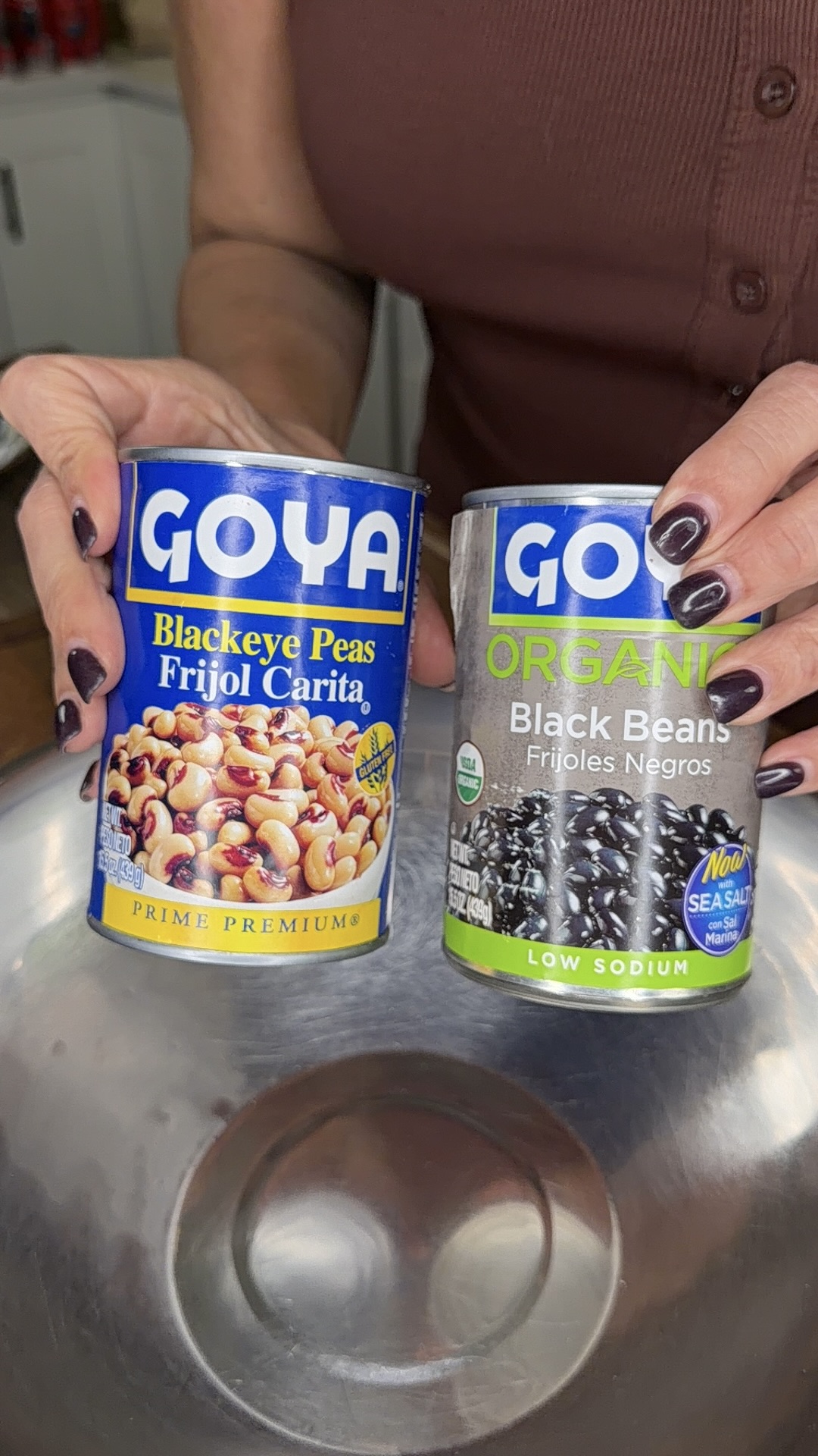 A person holds two cans: Goya Blackeye Peas (Frijol Carita) in the left hand and Goya Organic Black Beans (Frijoles Negros) in the right hand, positioned above a metal bowl.