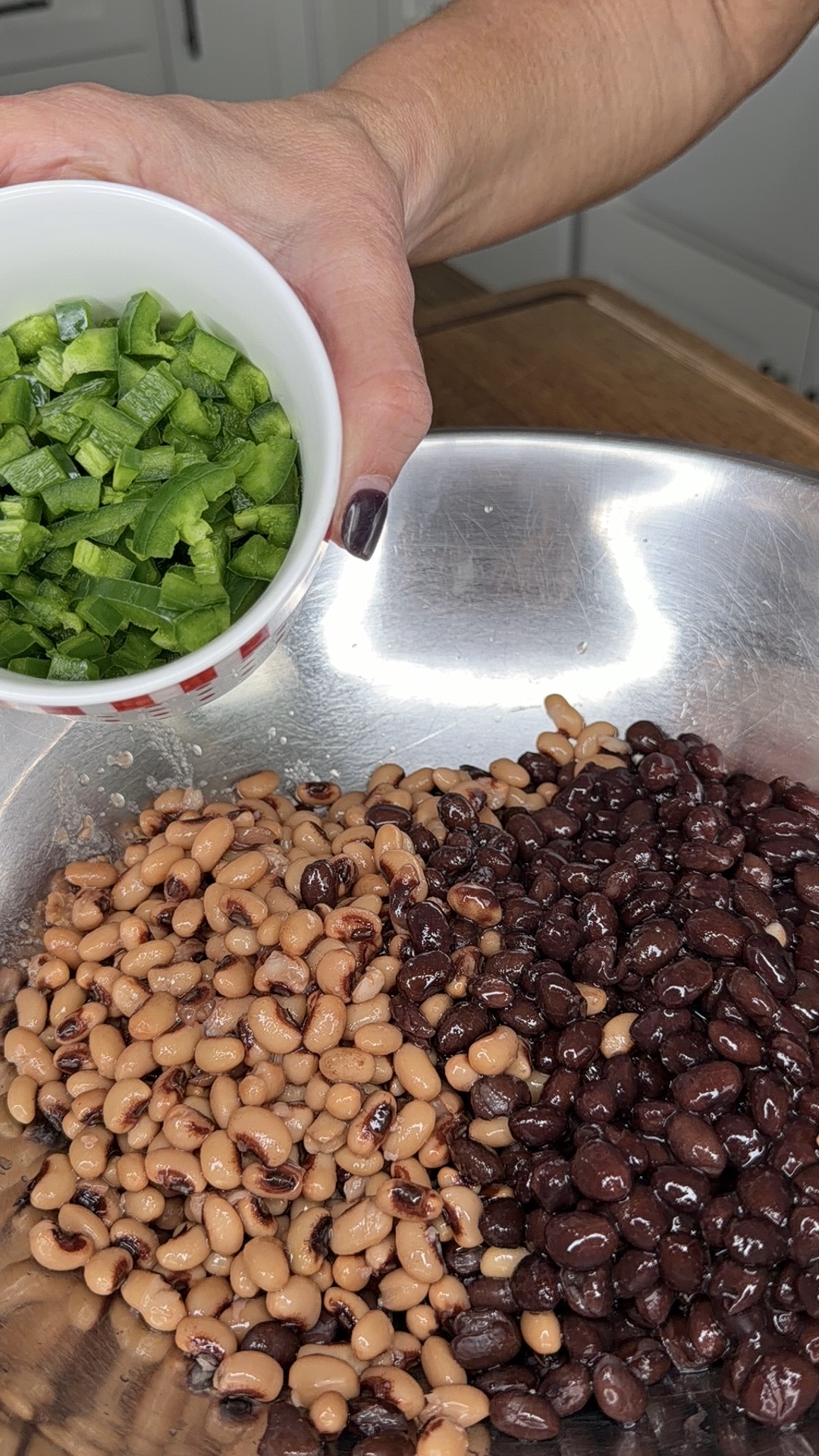 A hand holds a small bowl of chopped green bell peppers above a large metal bowl filled with black beans and black-eyed peas. Kitchen counters are visible in the background.