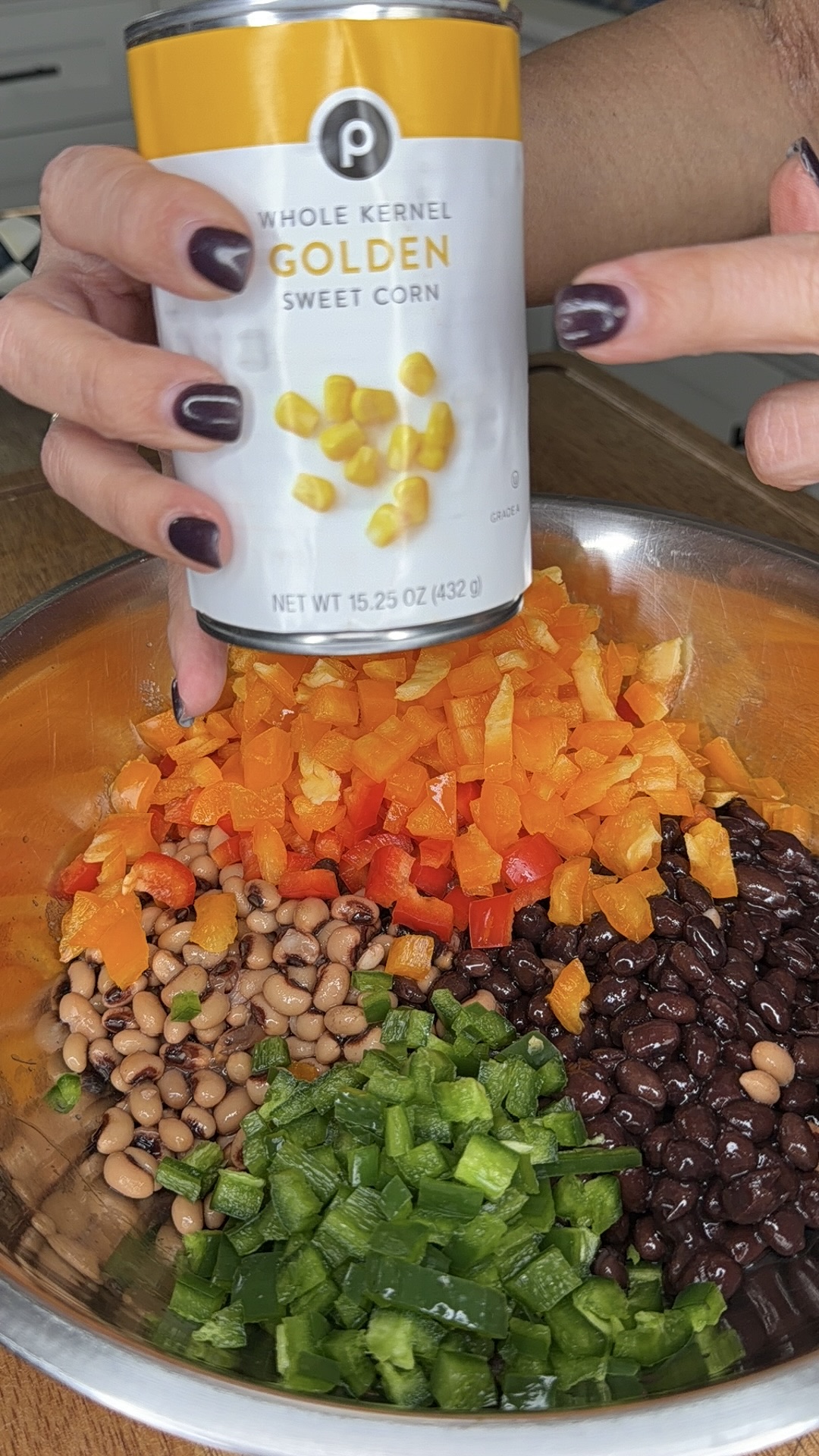 A person with dark nail polish holds a can of golden sweet corn above a bowl filled with chopped orange bell peppers, jalapeños, black beans, and black-eyed peas.