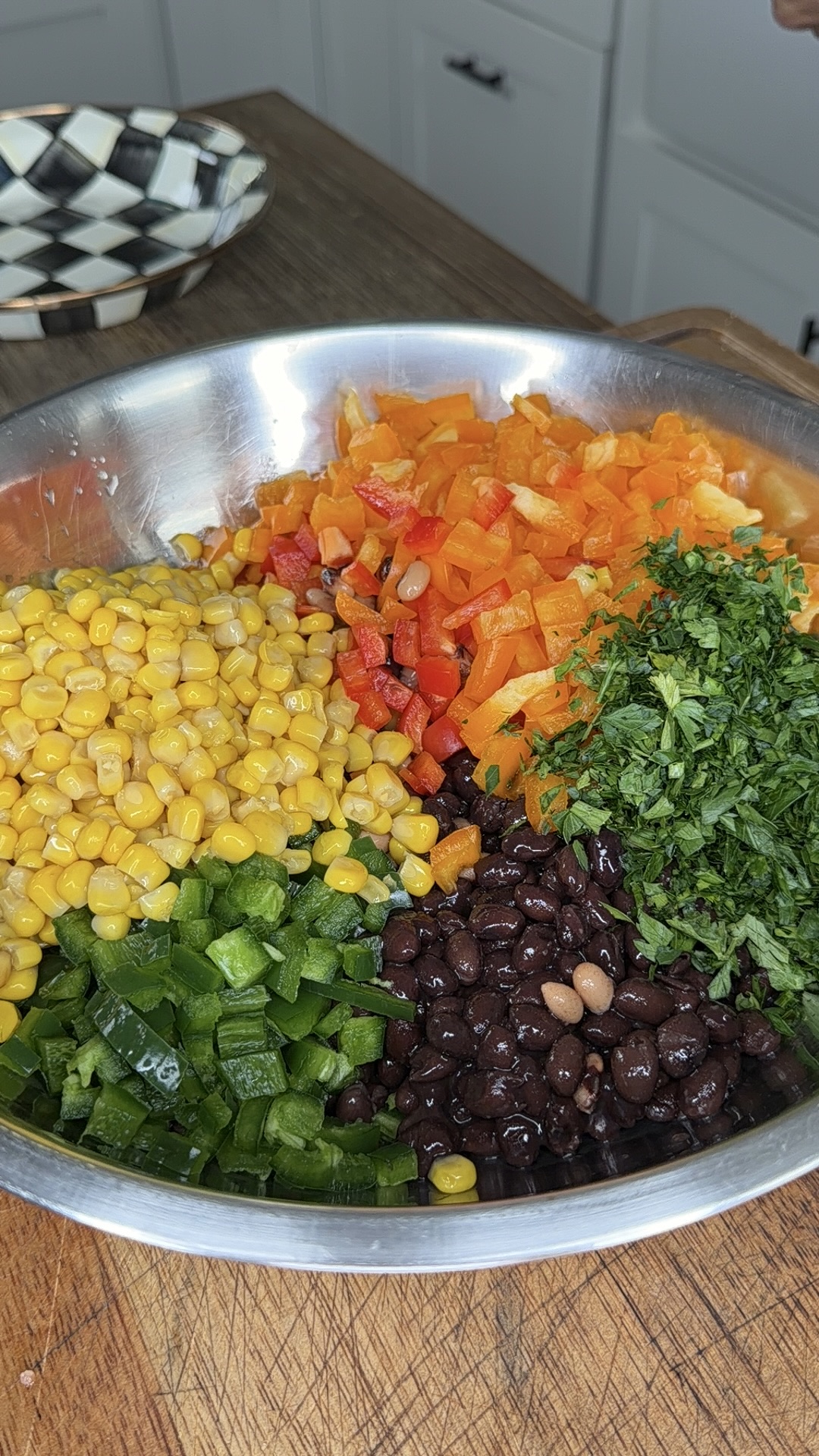 A metal bowl filled with chopped orange and red bell peppers, yellow corn, black beans, green jalapeños, and fresh cilantro, placed on a wooden counter in a kitchen.