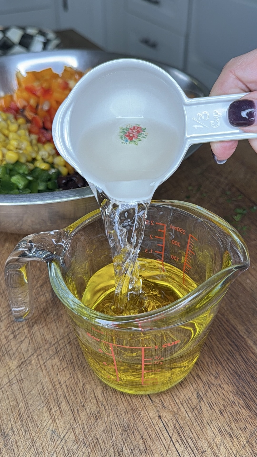 A hand pours clear liquid from a measuring cup into a larger glass measuring cup containing yellow oil. In the background, a bowl of chopped vegetables sits on a wooden surface.