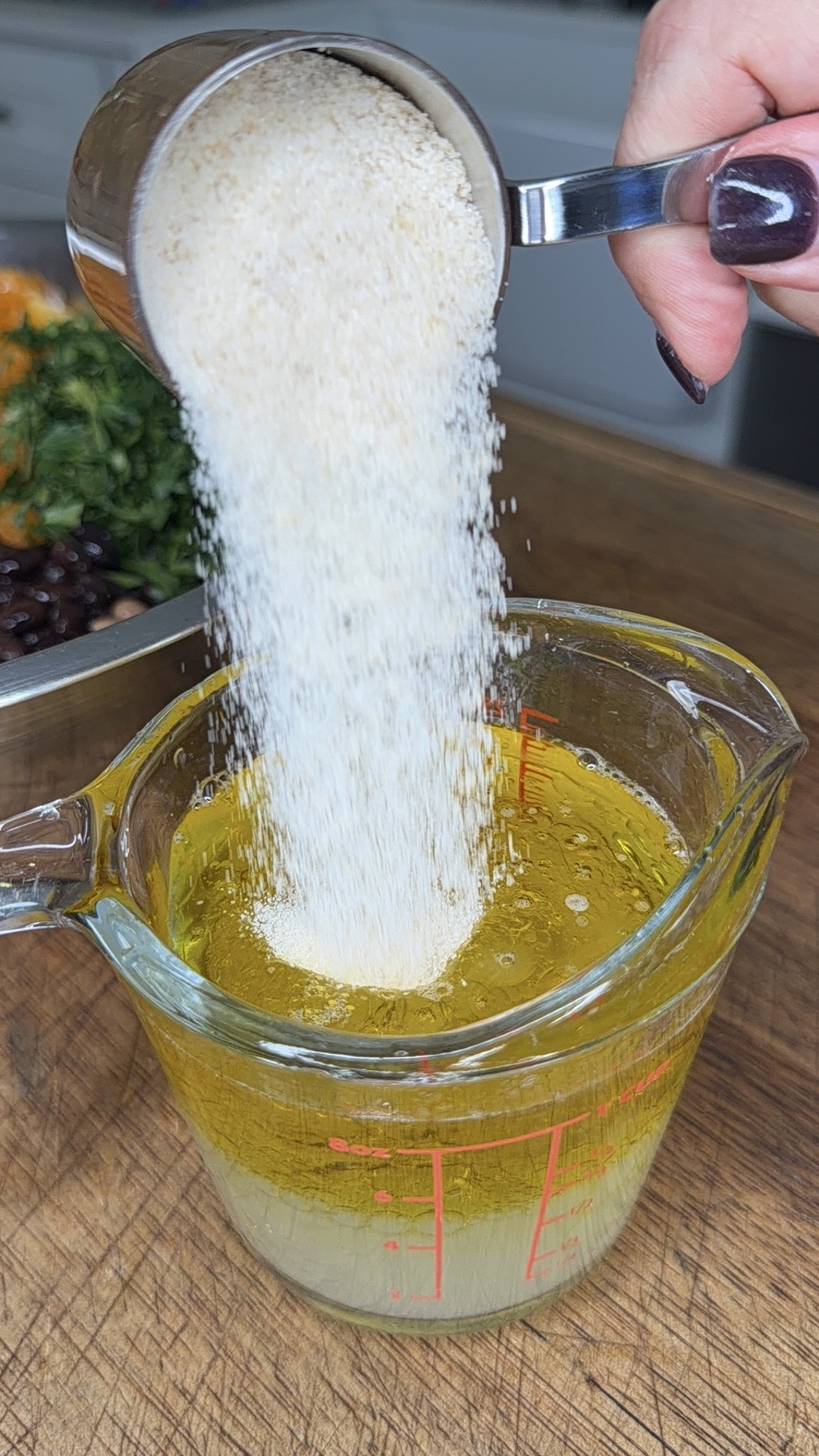 A hand pours a measuring cup of dry breadcrumbs into a glass measuring cup filled with olive oil on a wooden counter, with herbs and beans visible in the background.