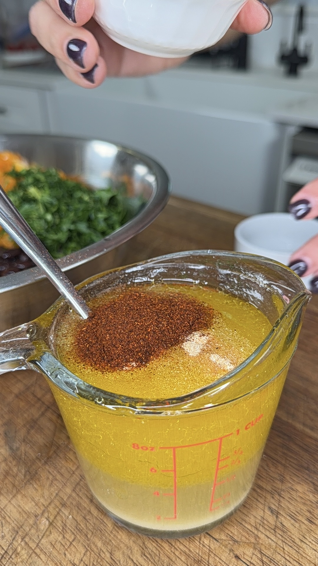A hand sprinkles spices into a glass measuring cup filled with yellow liquid, likely a dressing, while another hand holds the cup. A bowl of chopped ingredients is visible in the background on a wooden surface.