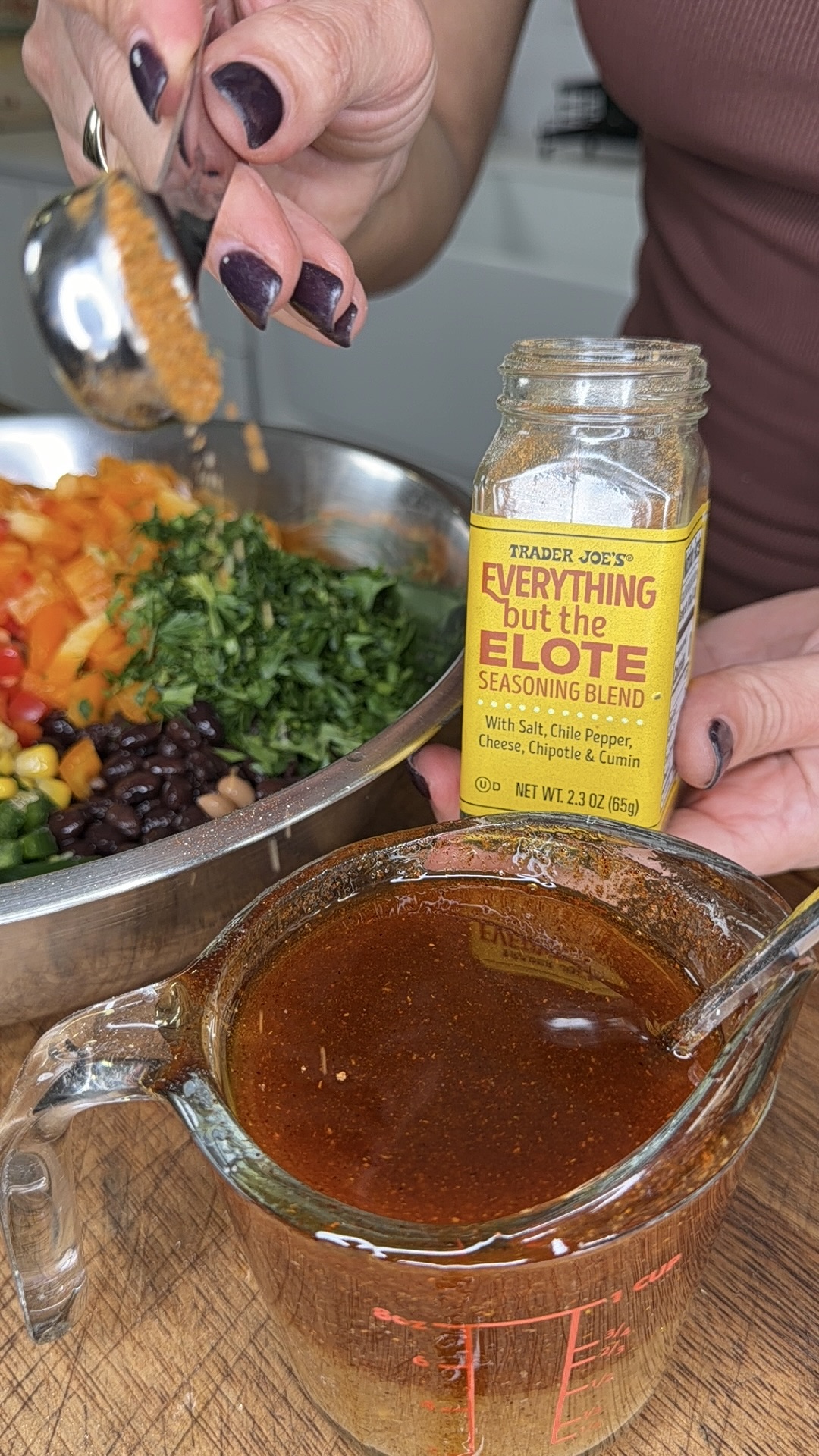 A person sprinkles Trader Joe’s Everything but the Elote seasoning blend from a jar into a bowl of chopped vegetables, with a glass measuring cup of sauce in the foreground.