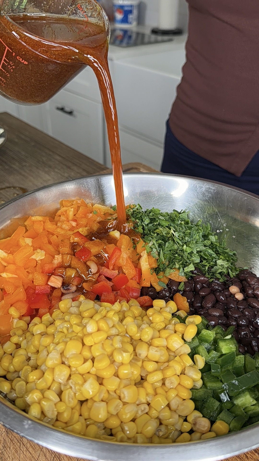 A person pours a reddish-brown dressing from a measuring cup into a bowl filled with corn, black beans, chopped bell peppers, cilantro, and jalapeños on a kitchen counter.
