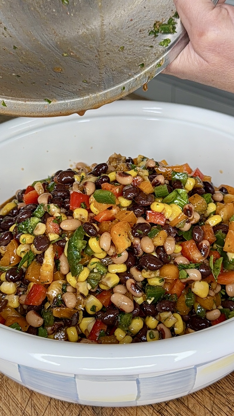 A close-up of a colorful bean salad with corn, black beans, chopped bell peppers, and herbs in a white bowl, as dressing is being poured from a metal pan onto the salad.