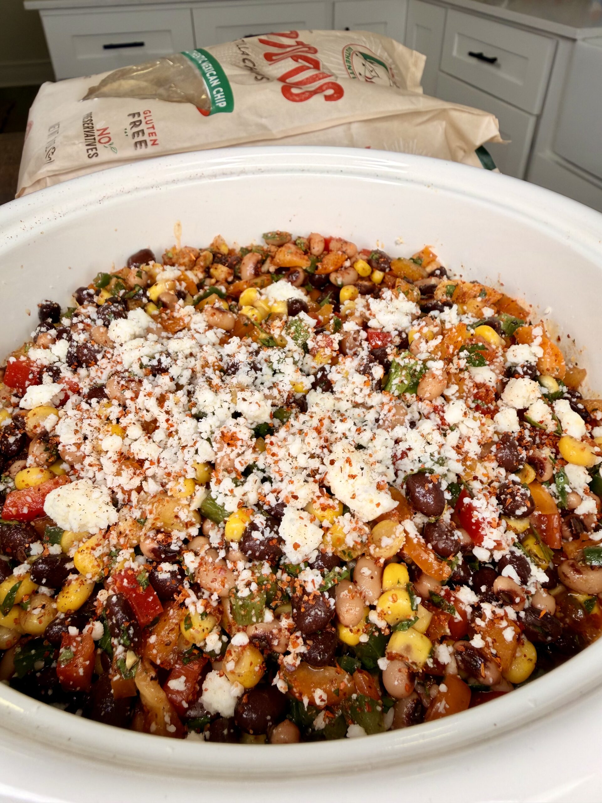 A bowl of colorful bean salad with black beans, corn, tomatoes, and crumbled cheese on top, sits on a counter with a bag of tortilla chips in the background.